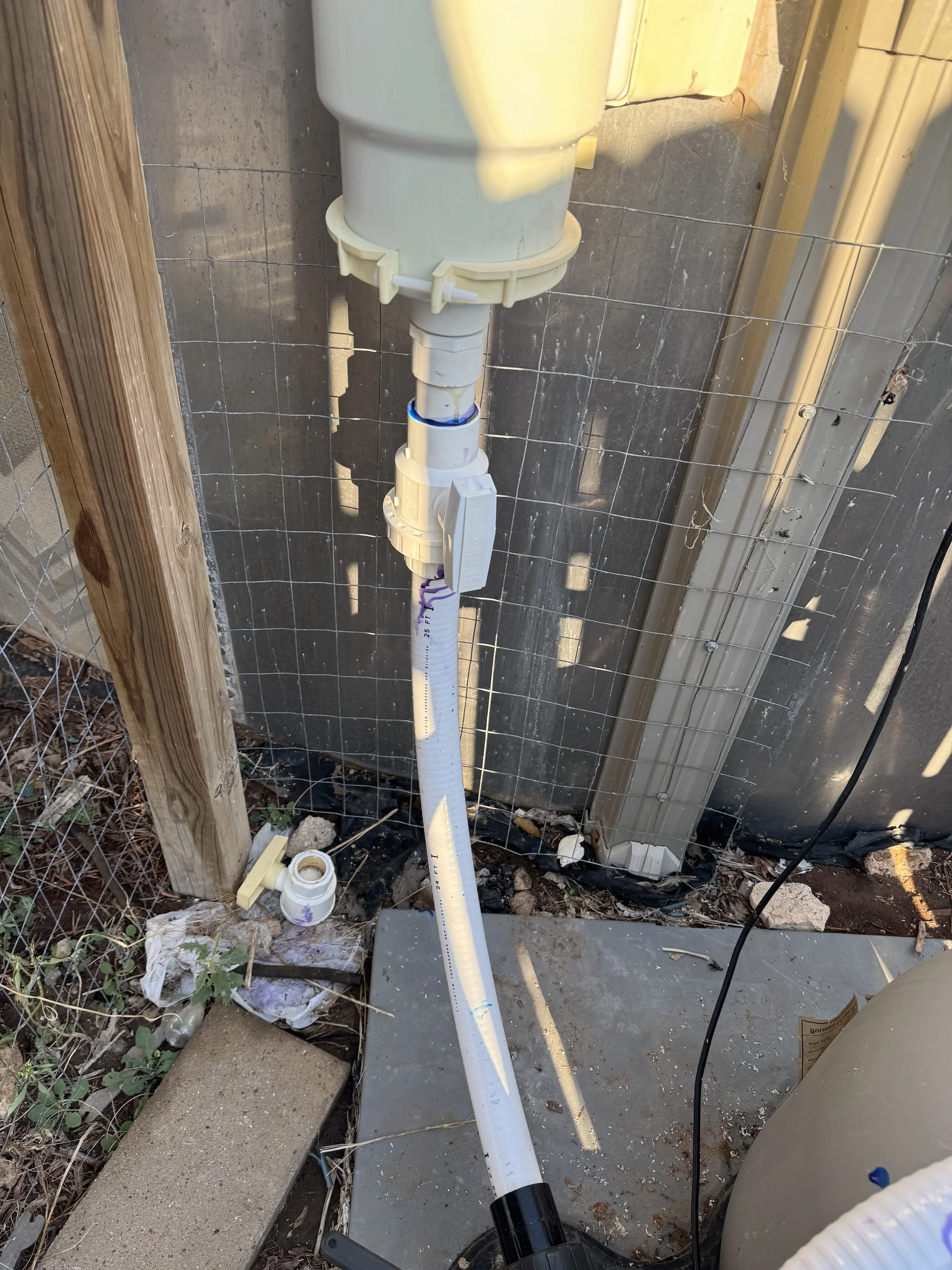 A white plastic pipe with a valve and fittings is installed vertically against a concrete wall, surrounded by a wire fence, with various construction debris and supplies on the ground.