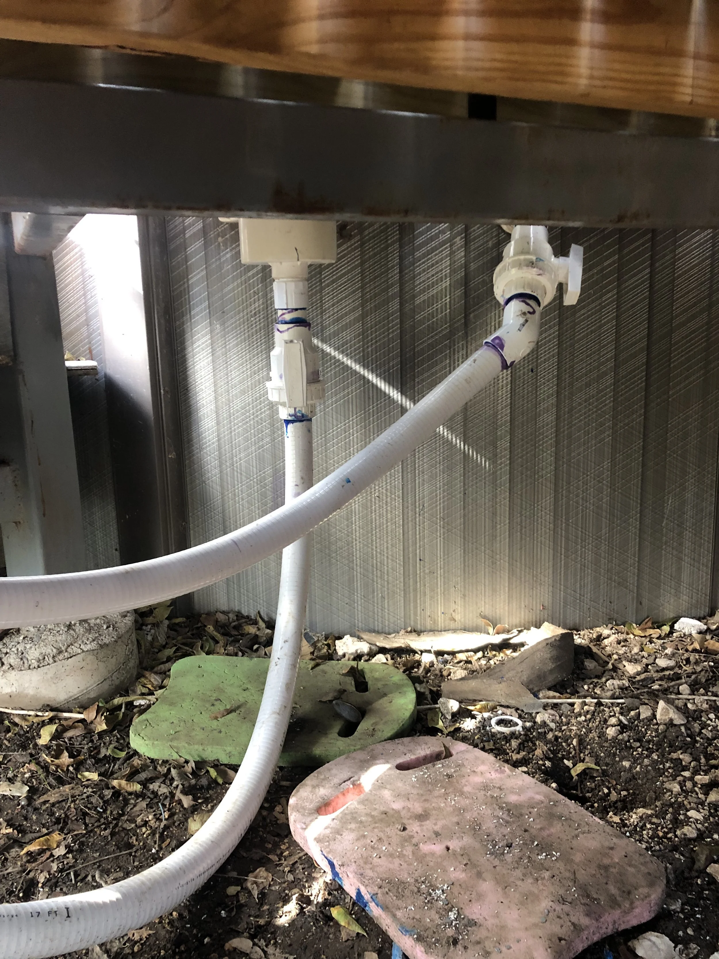 Underground plumbing pipes, including a white flexible hose, connected to a beige box fitting, mounted under a wooden structure, with a concrete and soil ground surface.