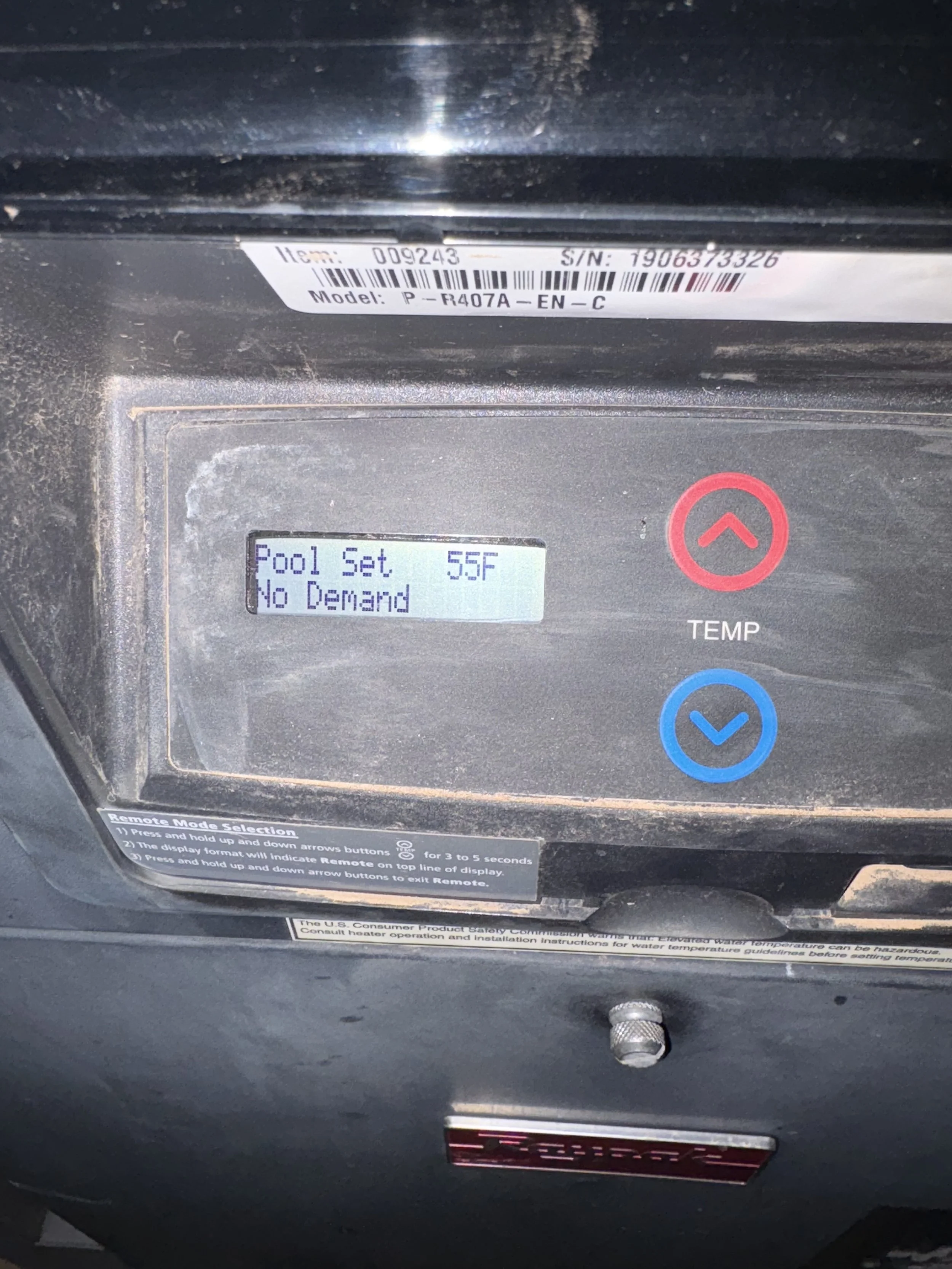 Close-up of a digital control panel for a water heater or pool heater with a small display showing temperature and settings, including 'Pool Set 55F' and 'No Demand', with red and blue arrow buttons, and a label with item, serial number, and model details.