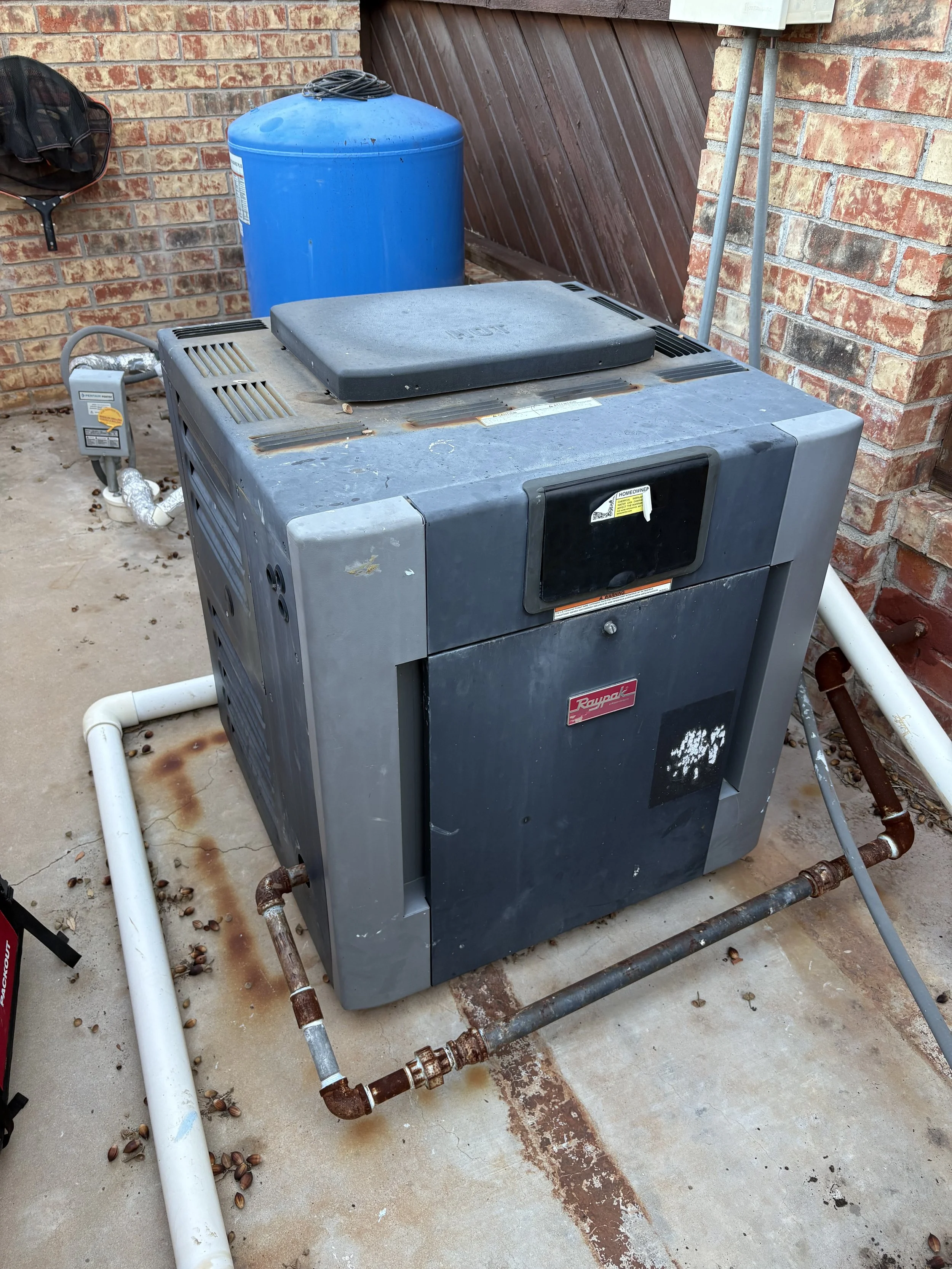 An outdoor HVAC unit with central piping, placed on a concrete ground, with a large blue water container, a brick wall, and a wooden door in the background.