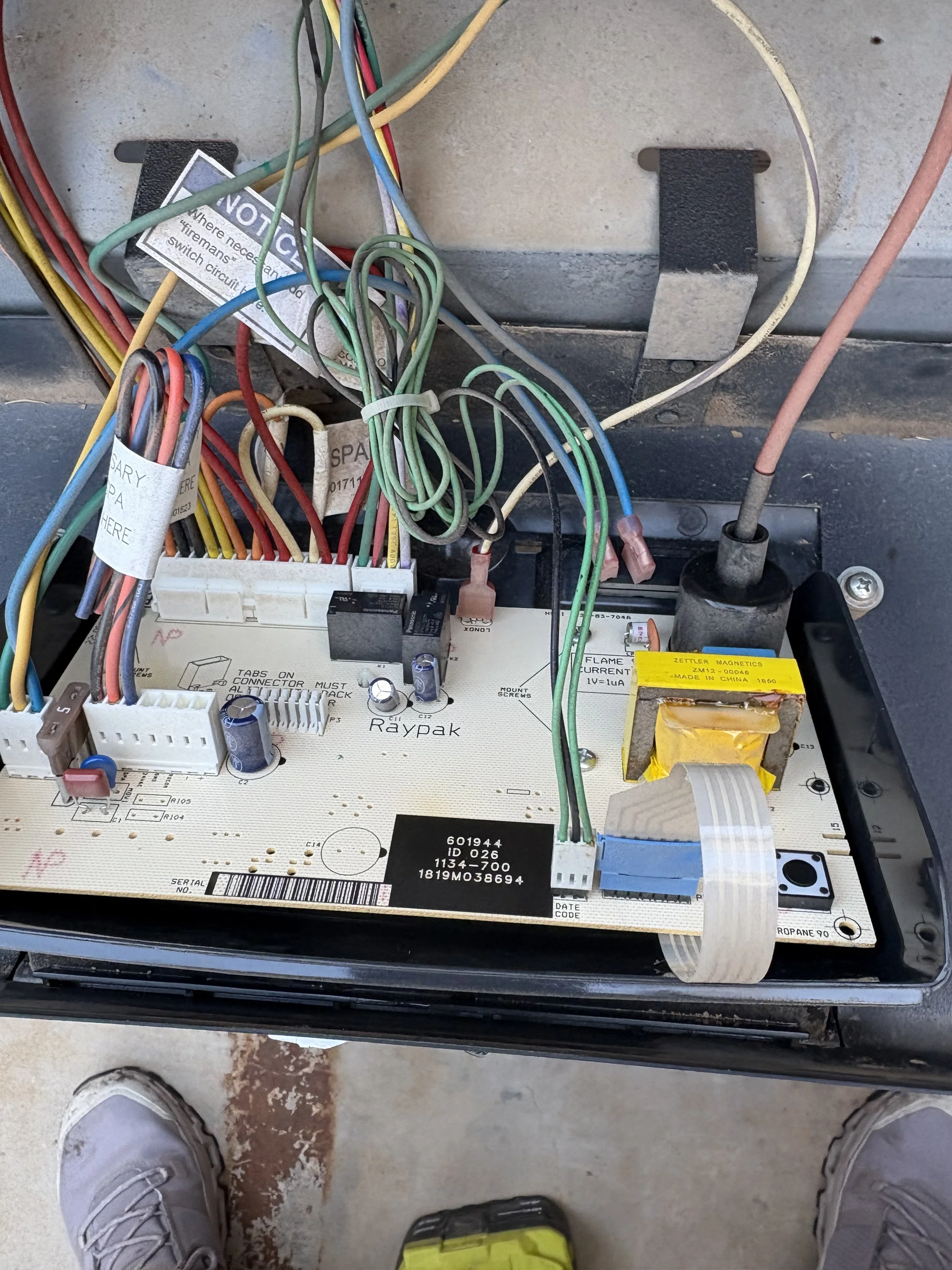 A person wearing gray sneakers and mustard yellow pants standing on a concrete floor in front of an open electrical panel with various colorful wires and circuit components inside.