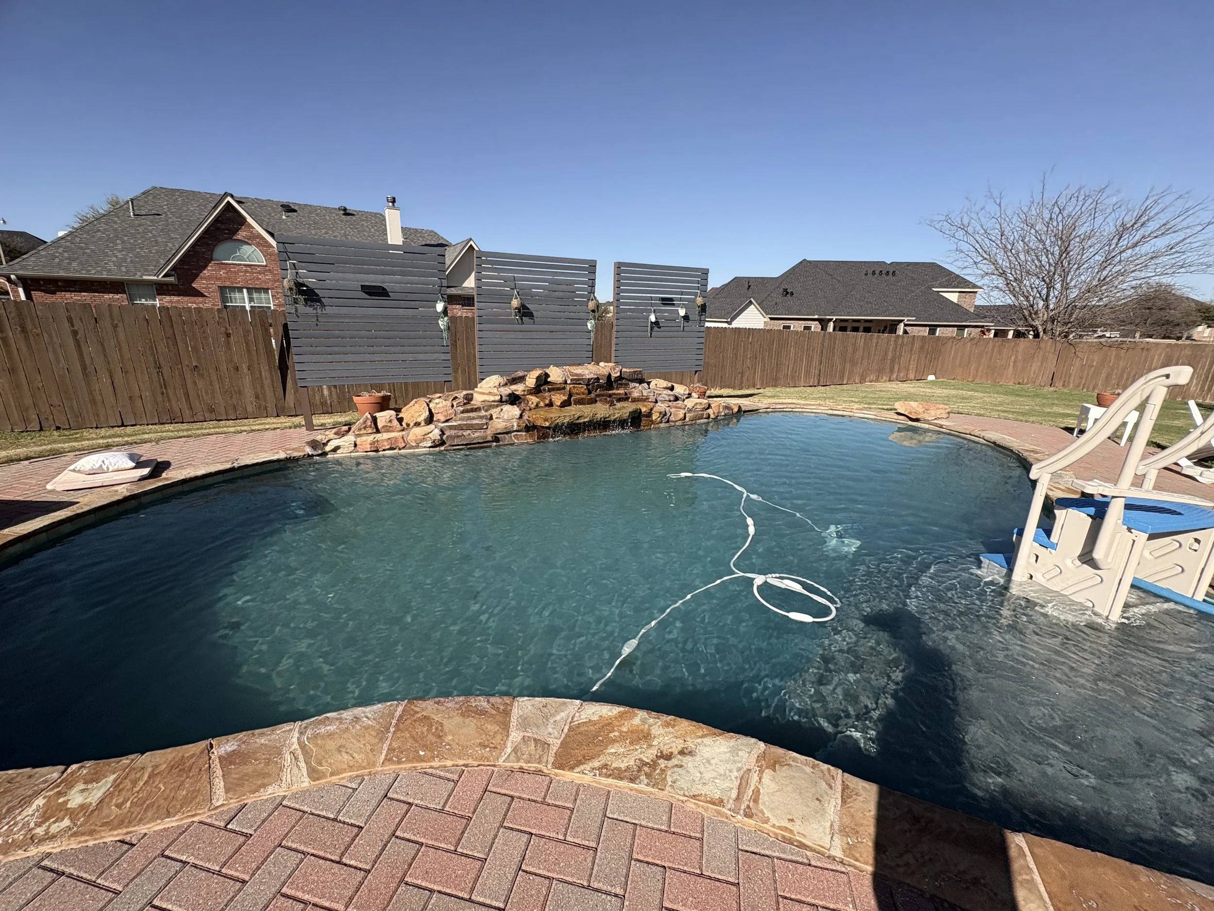 Backyard with swimming pool, rock waterfall feature, wooden privacy fence, and neighboring houses with sloped roofs.
