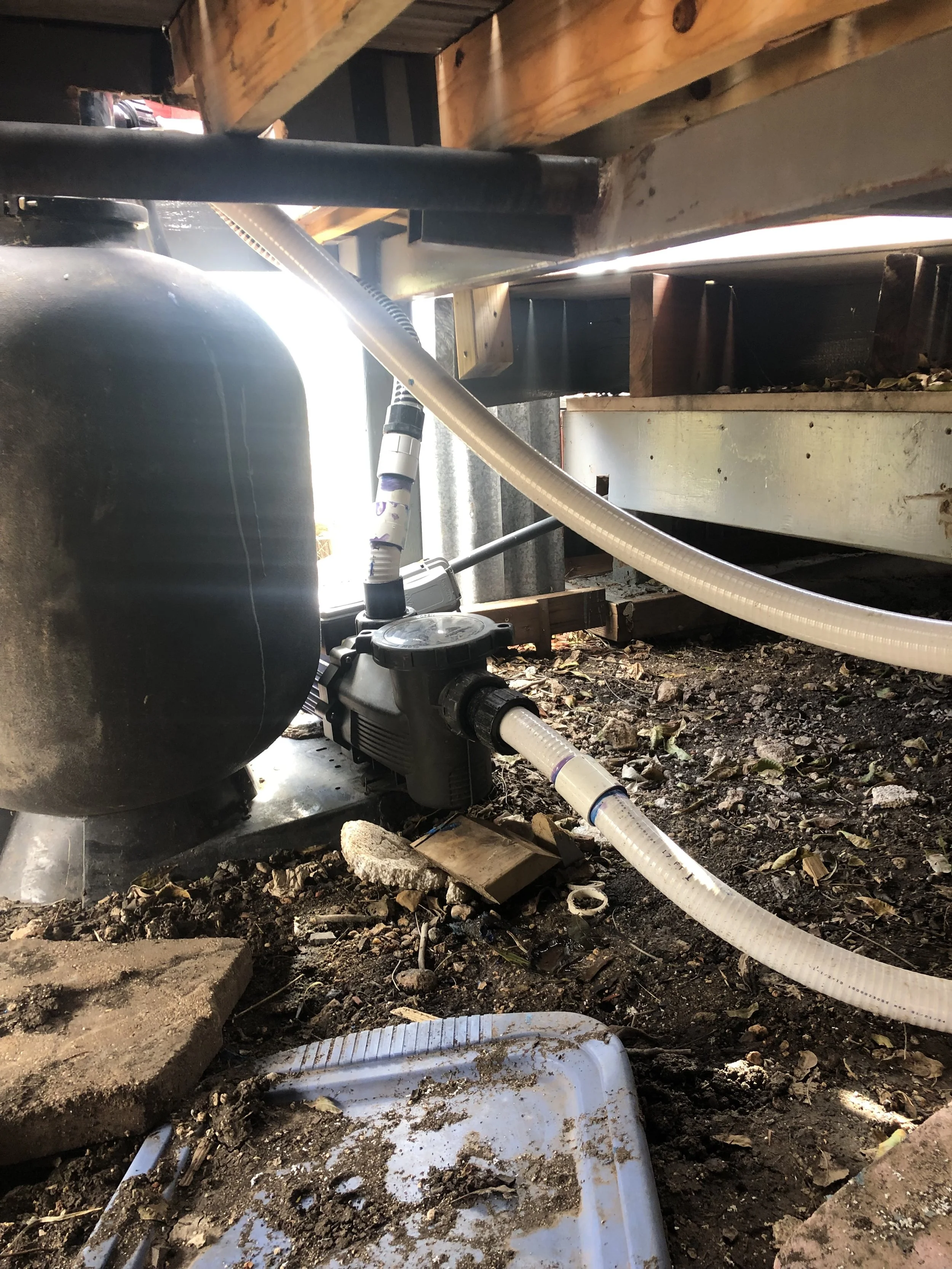 Underneath a structure, showing pipes, wires, and other plumbing equipment on a dirt ground with debris and leaves scattered around.
