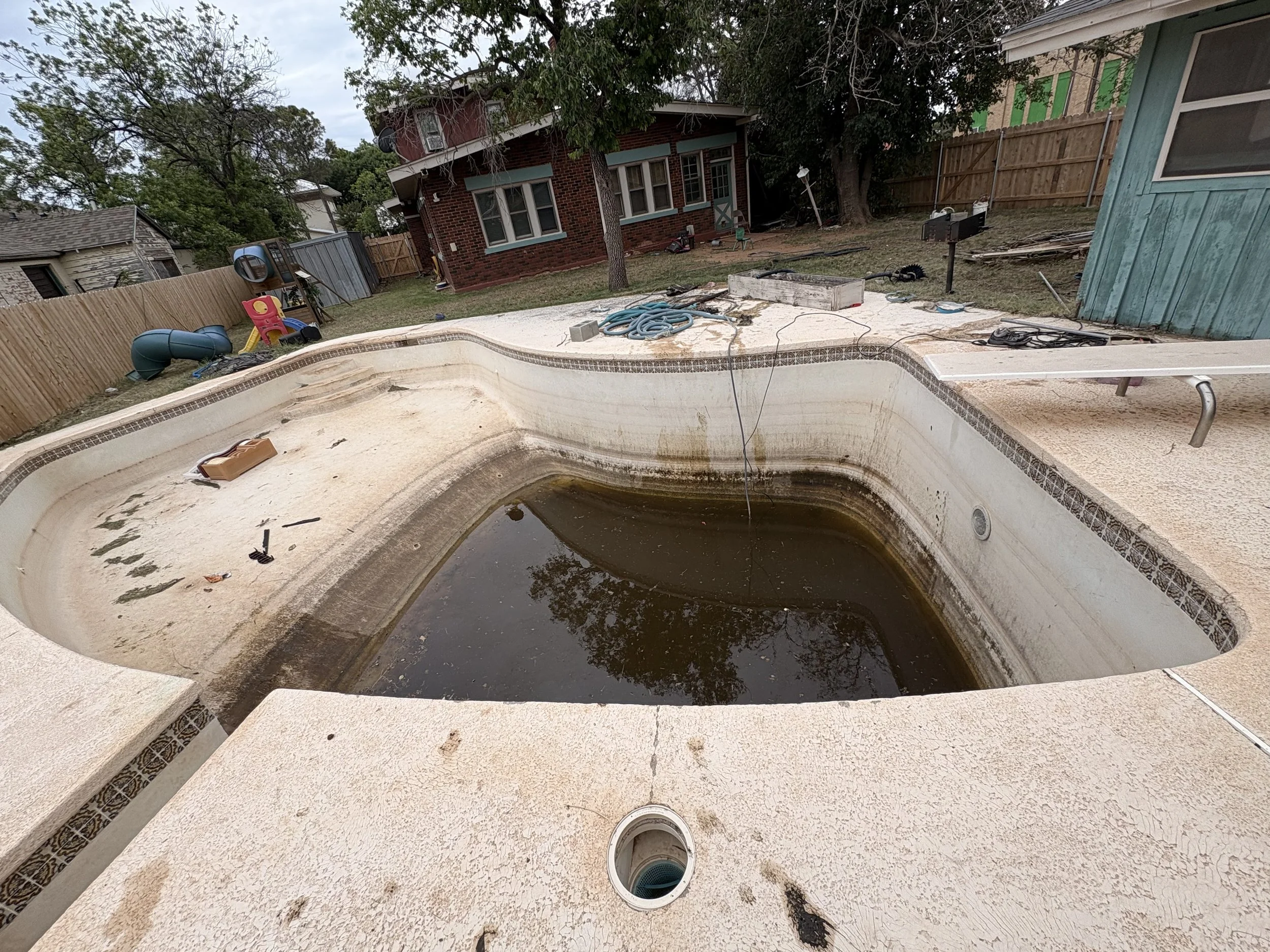 Empty swimming pool with dirty water and debris, surrounded by a concrete deck, with a pool skimmer and hose on the ground, and a backyard with trees and houses in the background.