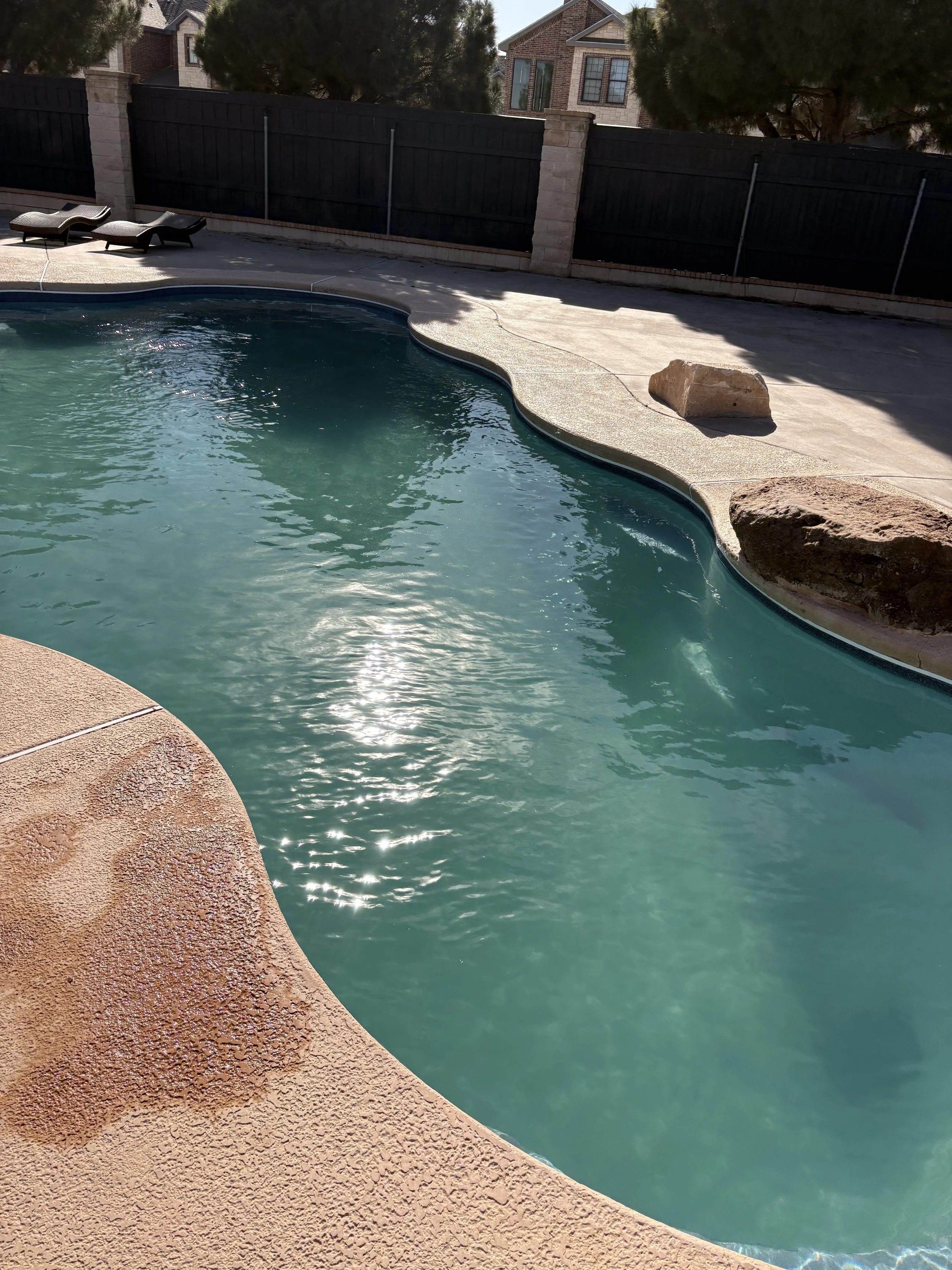 An outdoor swimming pool with clear blue water, surrounded by a textured pinkish concrete deck and a black fence, with some residential houses and trees visible in the background.