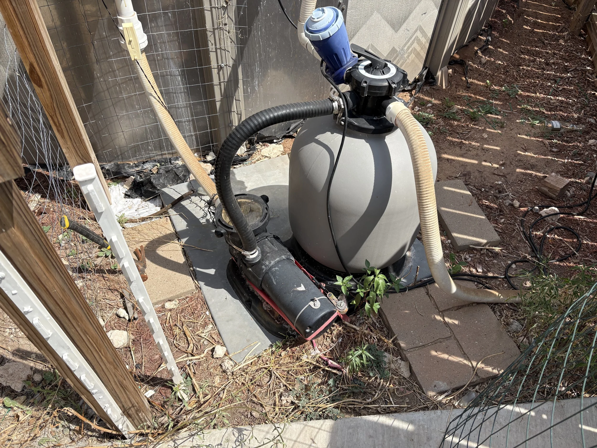 A pool filter system with hoses connected, situated outdoors on a concrete pad, next to wooden fencing and a wire mesh panel, with dirt, small plants, and an empty can nearby.
