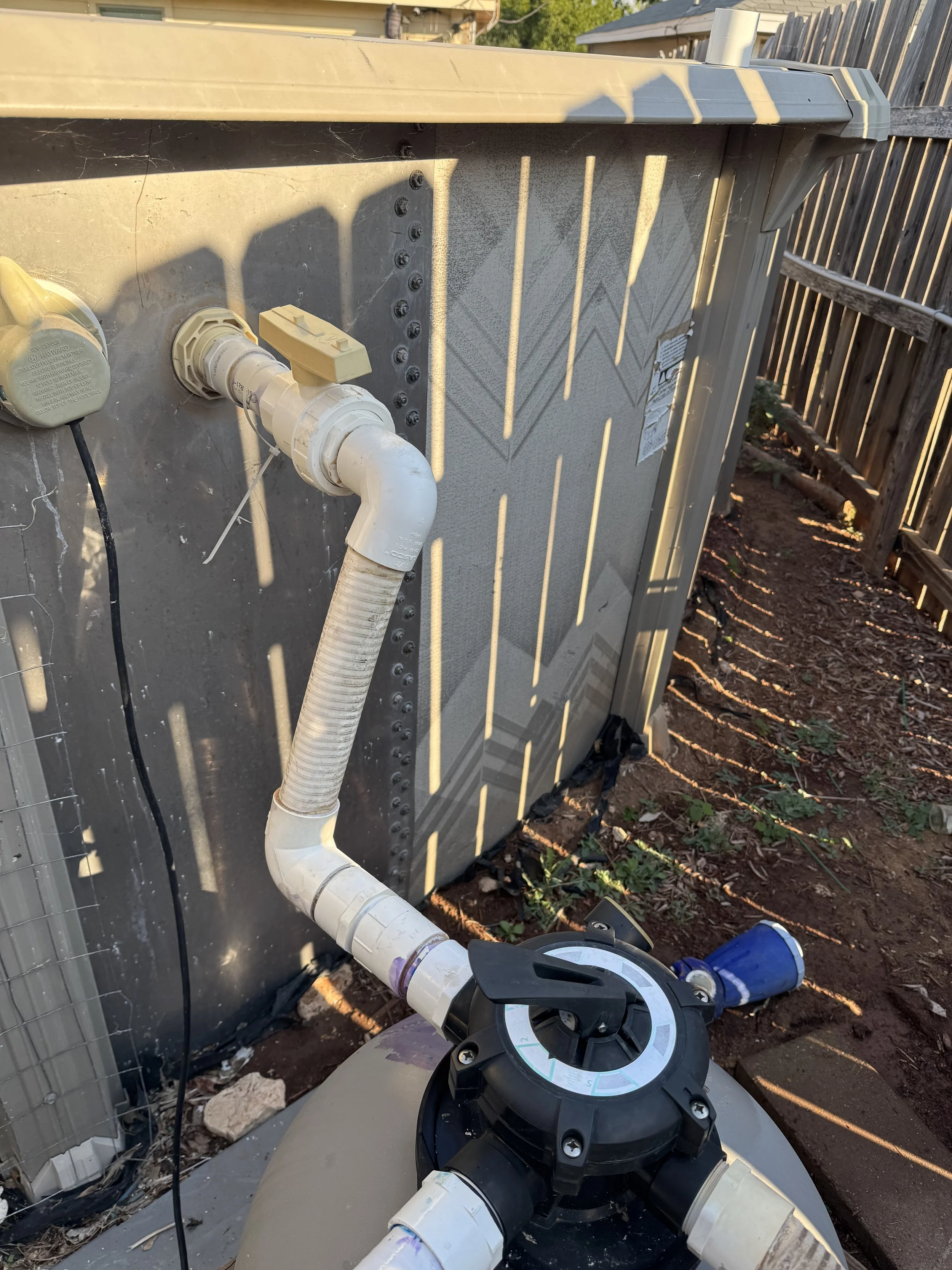 An outdoor view of a swimming pool pump and piping system, with a backyard fence in the background and sunlight casting shadows on the equipment.