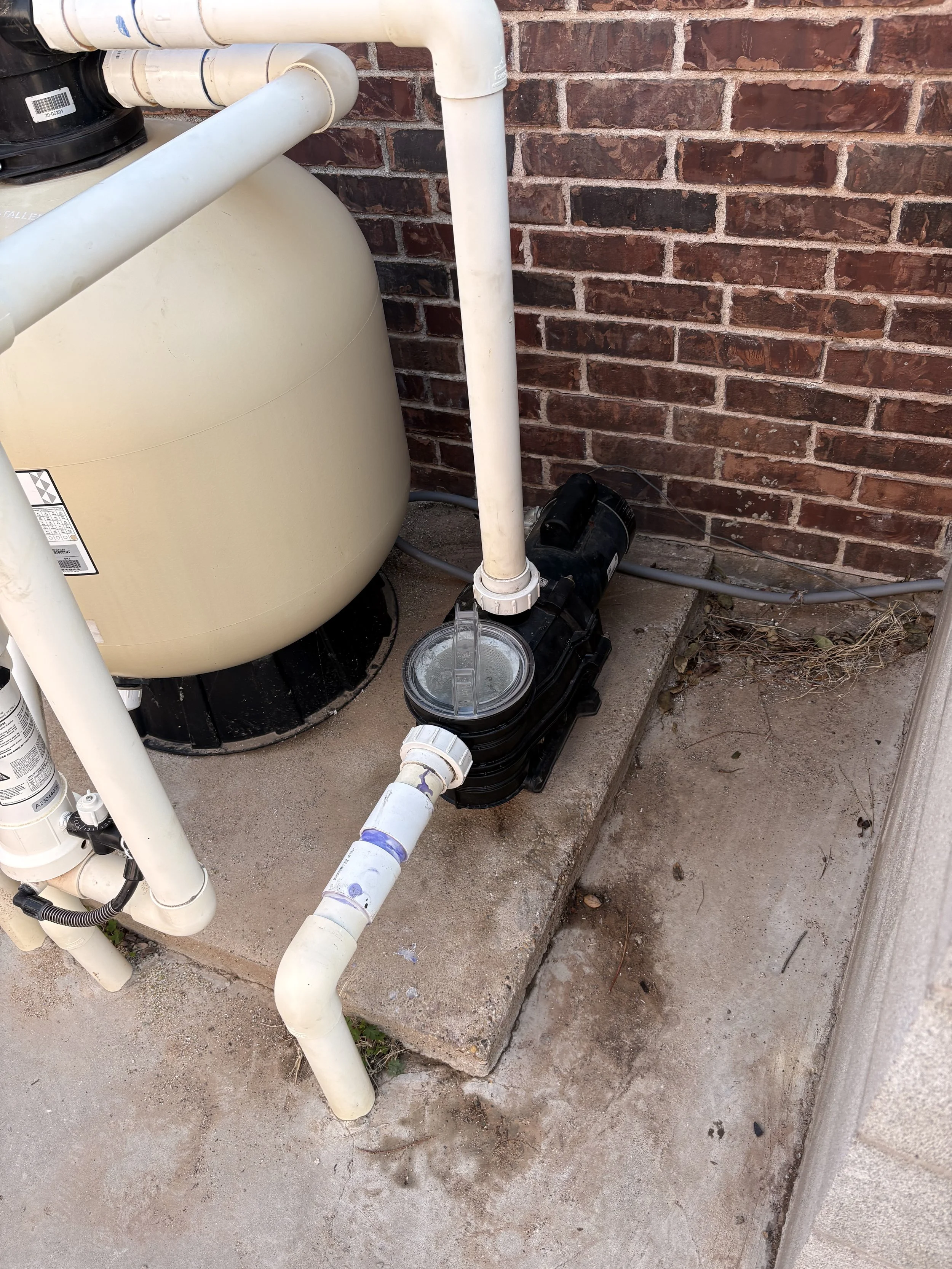 Close-up of a water filtration or pump system installed outdoors against a brick wall, with white PVC pipes, a large beige tank, and a black motor at the base, with some dirt and debris on a concrete surface.