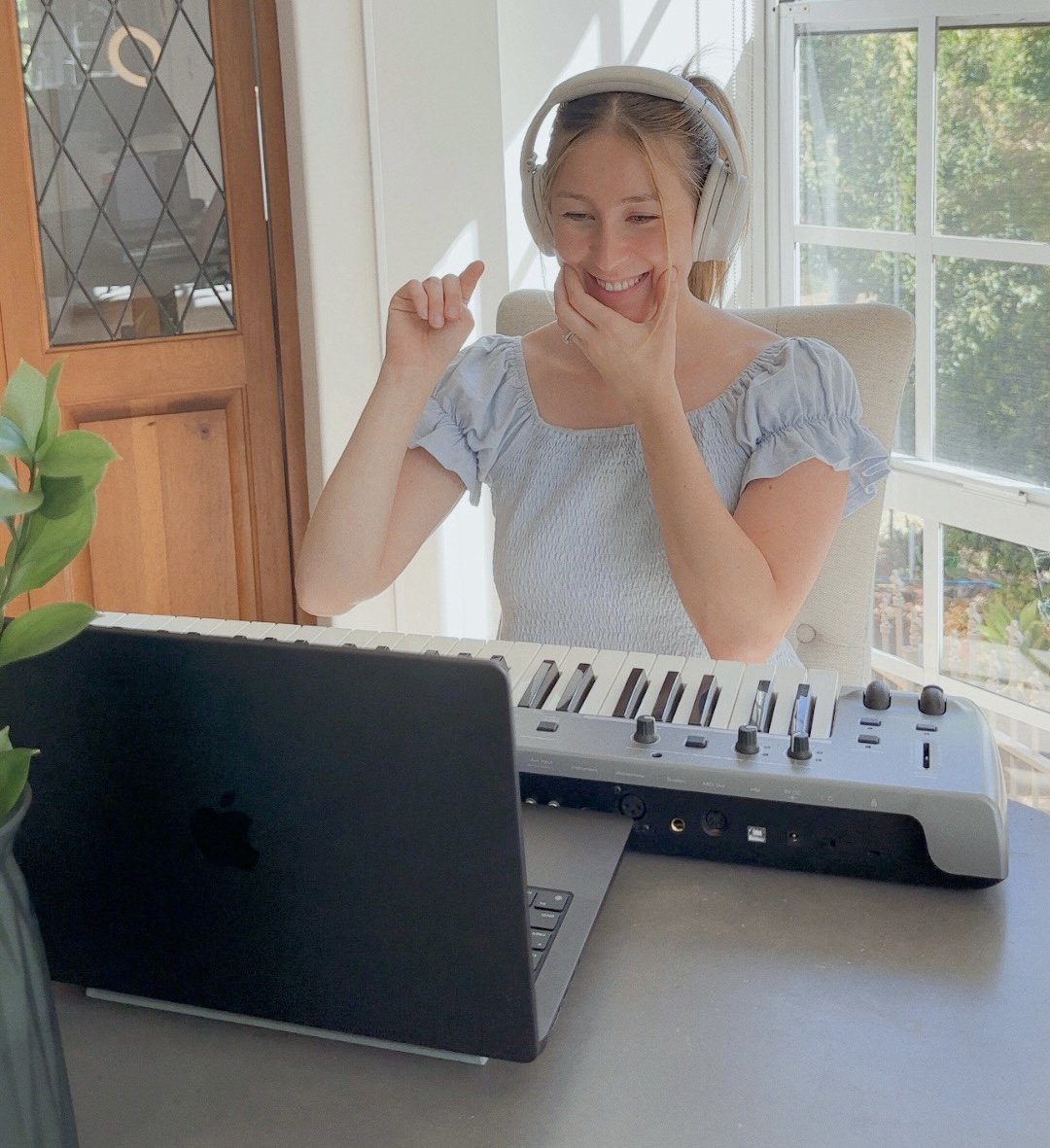 A woman wearing headphones, sitting at a desk with a keyboard, smiling while looking at a laptop screen running an online singing lesson