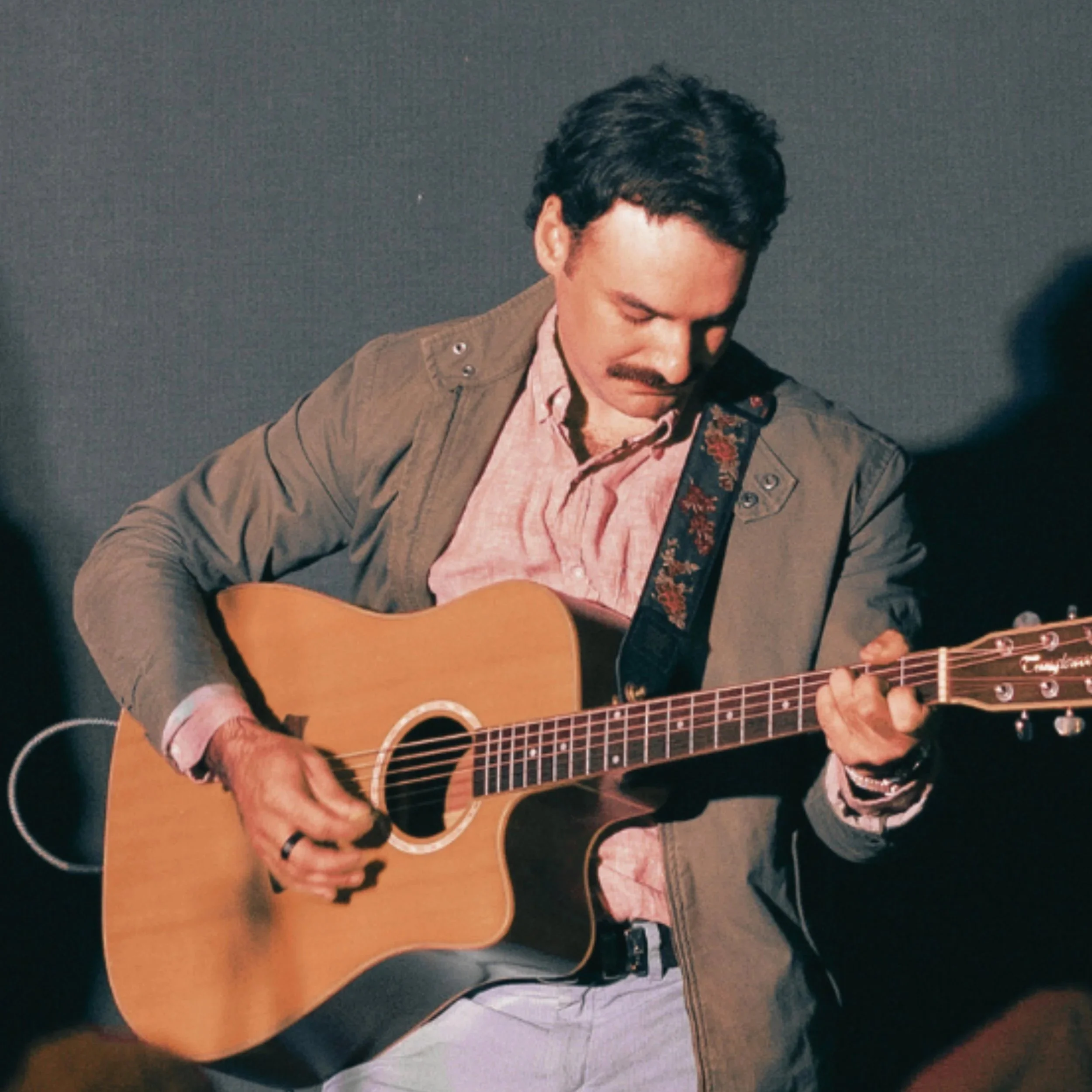 A man with dark hair and a mustache playing an acoustic guitar on stage.