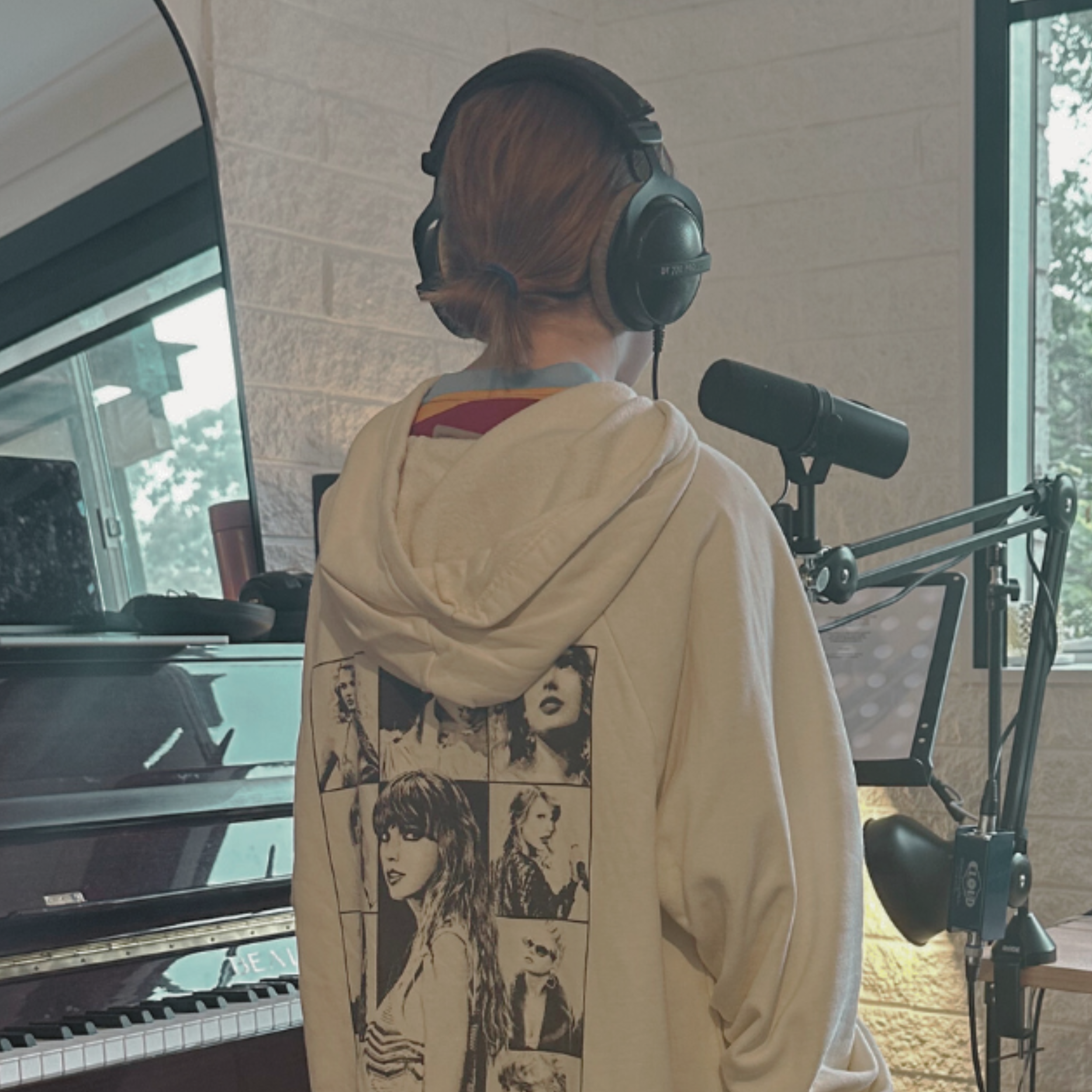 A person with reddish hair tied back, wearing headphones, stands in front of a microphone wearing a cream hoodie with black-and-white cartoon images on the back, in a room with a window and a piano. Doing a recording session
