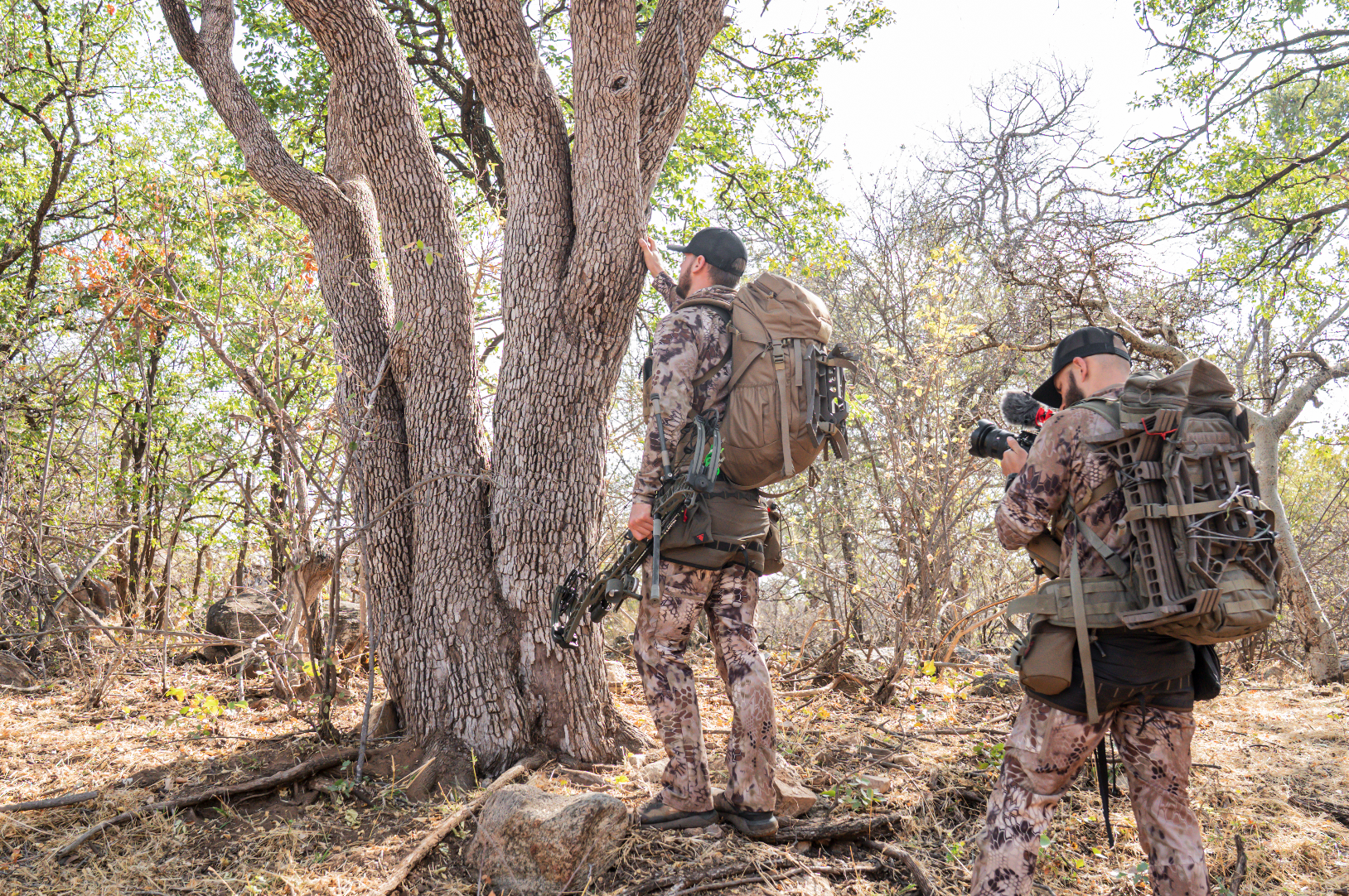Two hunters in camouflage clothing and backpacks are in a wooded area, one is reaching up towards a tree while the other is taking pictures of the scene.