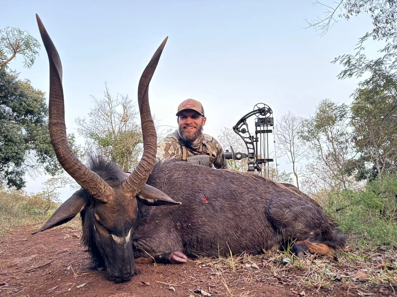 A smiling man in camouflage clothing sitting behind a large dead waterbuck with long, curved horns on the ground in a wooded area.