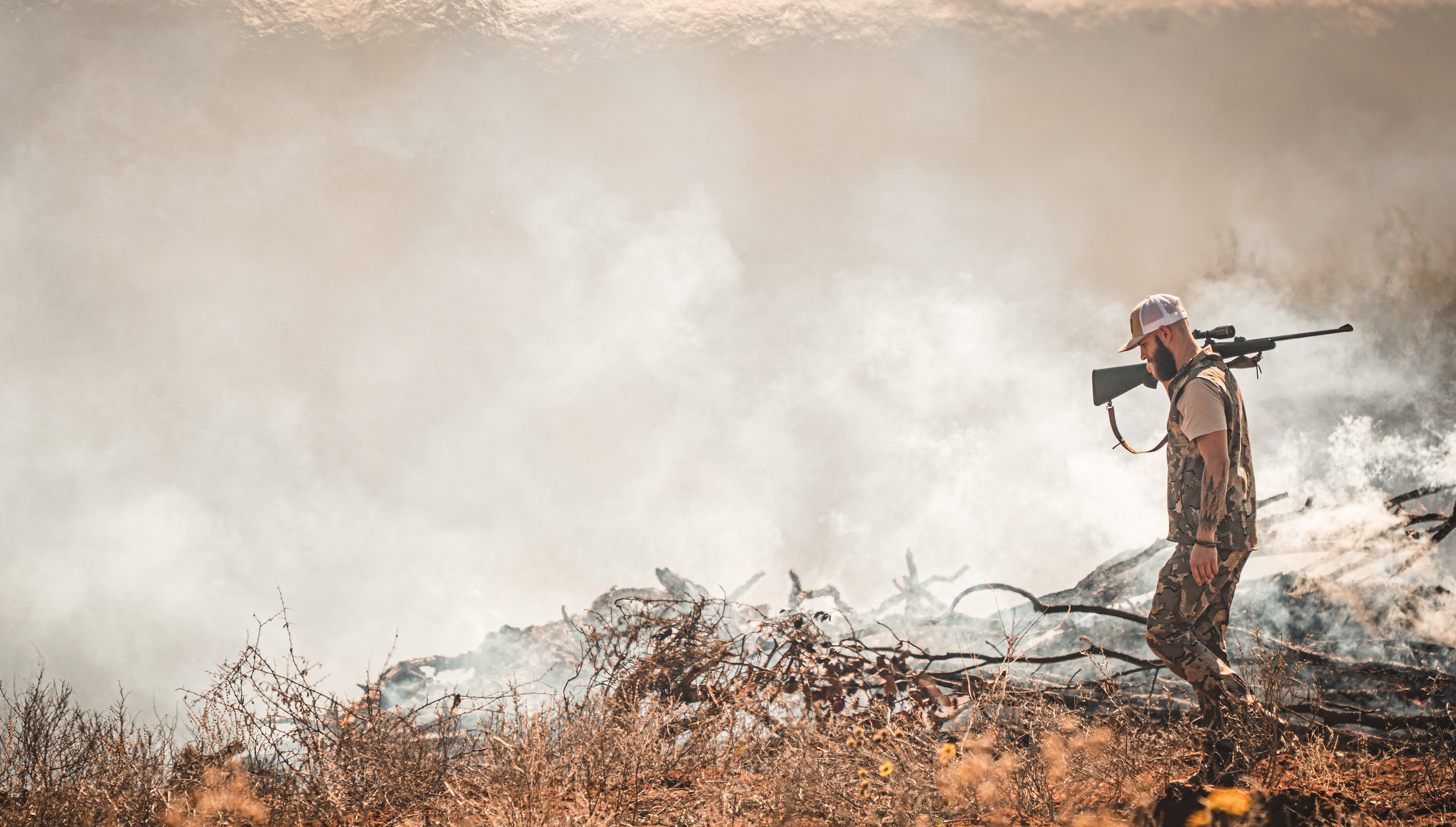 A man in camouflage clothing and a white cap carries a rifle while walking through a smoky, burn-damaged area outdoors during a wildfire.