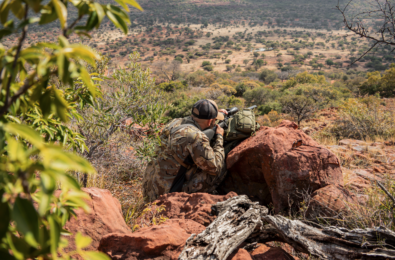 A person in camouflage clothing aiming a rifle from behind large red rocks in a semi-arid landscape with sparse trees and bushes.