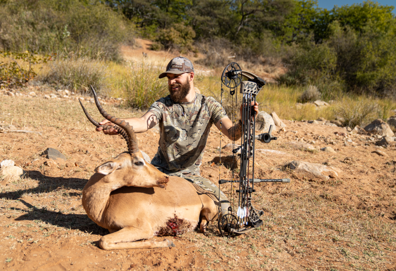 A man in camouflage clothing is sitting in the desert, holding a bow next to a recently hunted antelope with large curved horns and a wound on its side.
