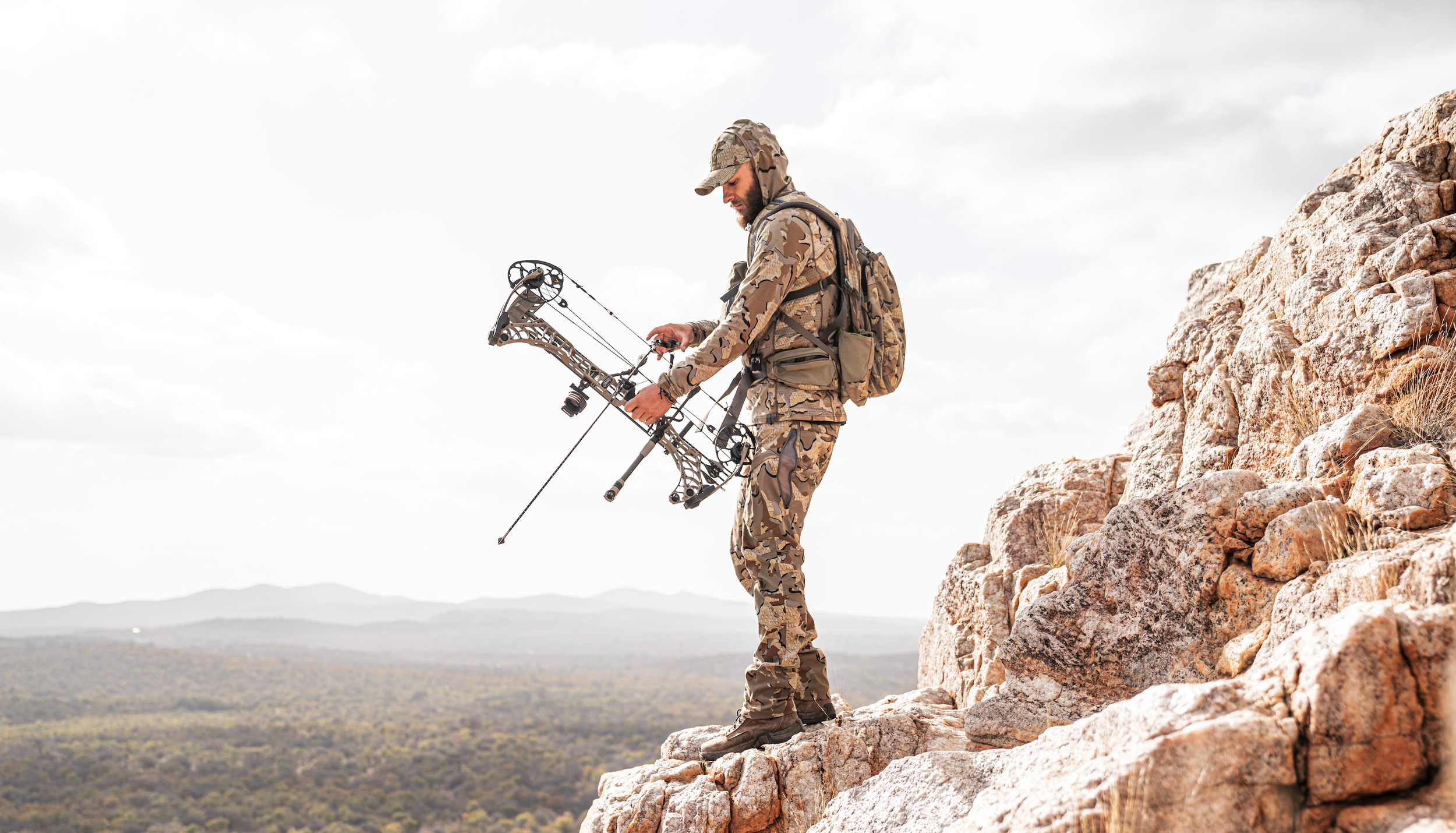 A man dressed in camouflage clothing stands on rocky terrain, holding a bow, ready to shoot an arrow, with a landscape of mountains and a cloudy sky in the background.