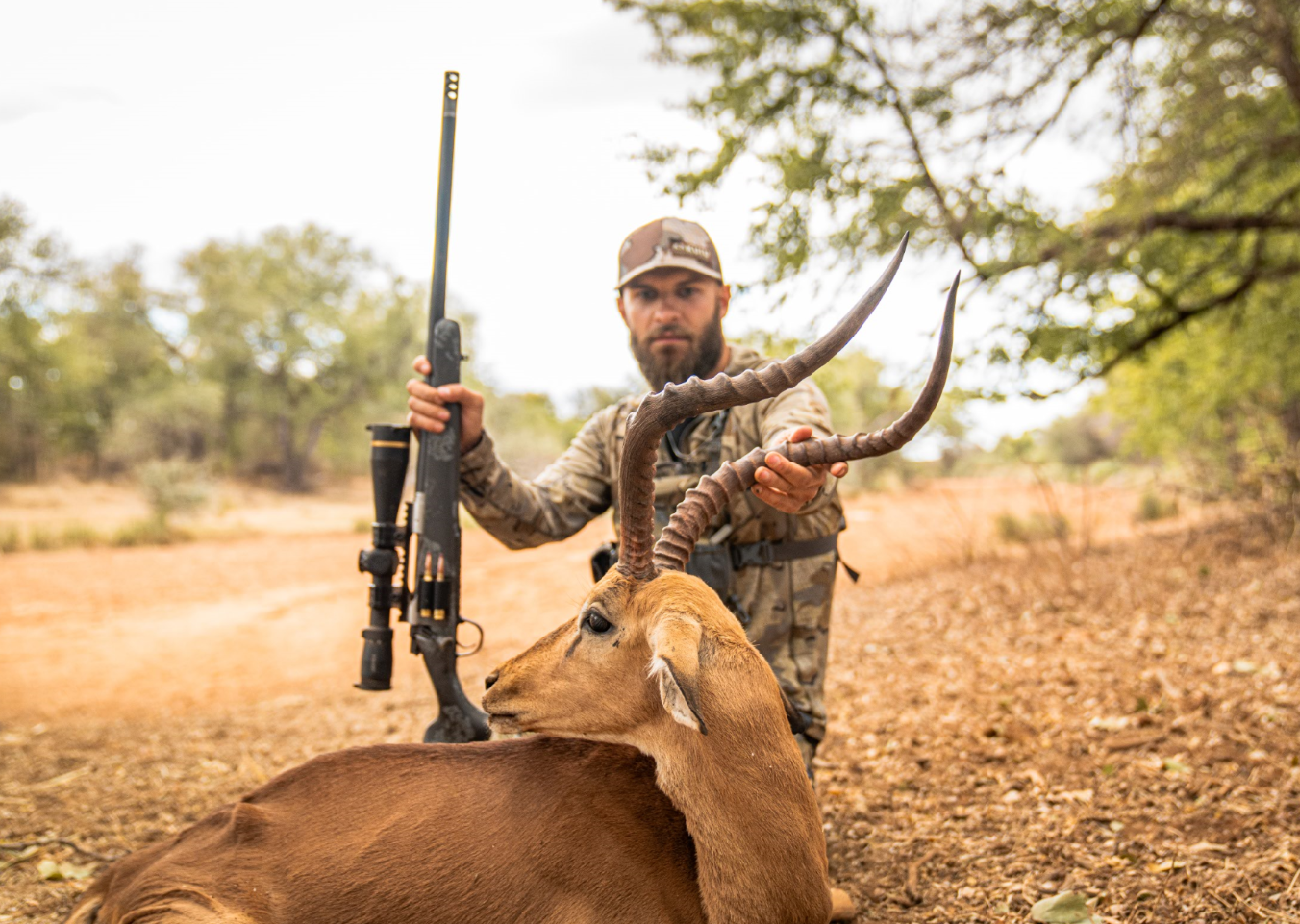 A man in camouflage clothes holding a hunting rifle behind a recently hunted antelope with curved horns, resting on the ground in a dry, wooded area.