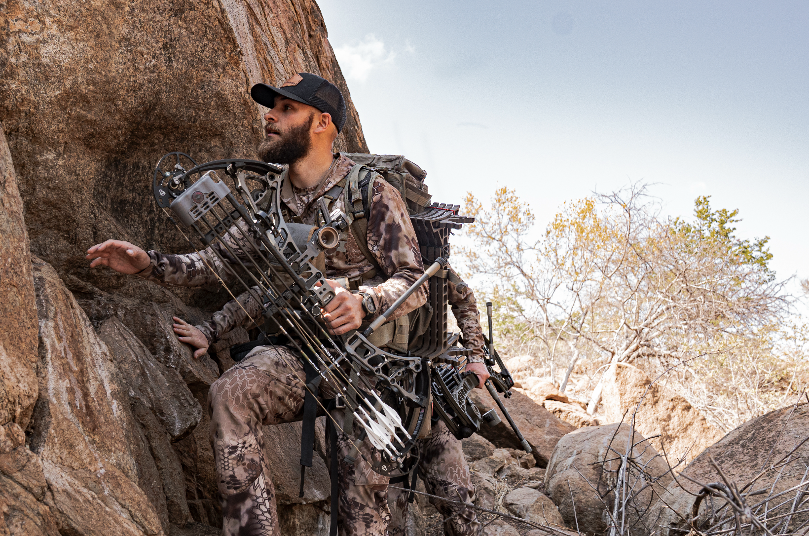 A man in camouflage climbing gear and a black cap holding a compound bow, positioned against a rocky outdoor terrain with trees in the background.