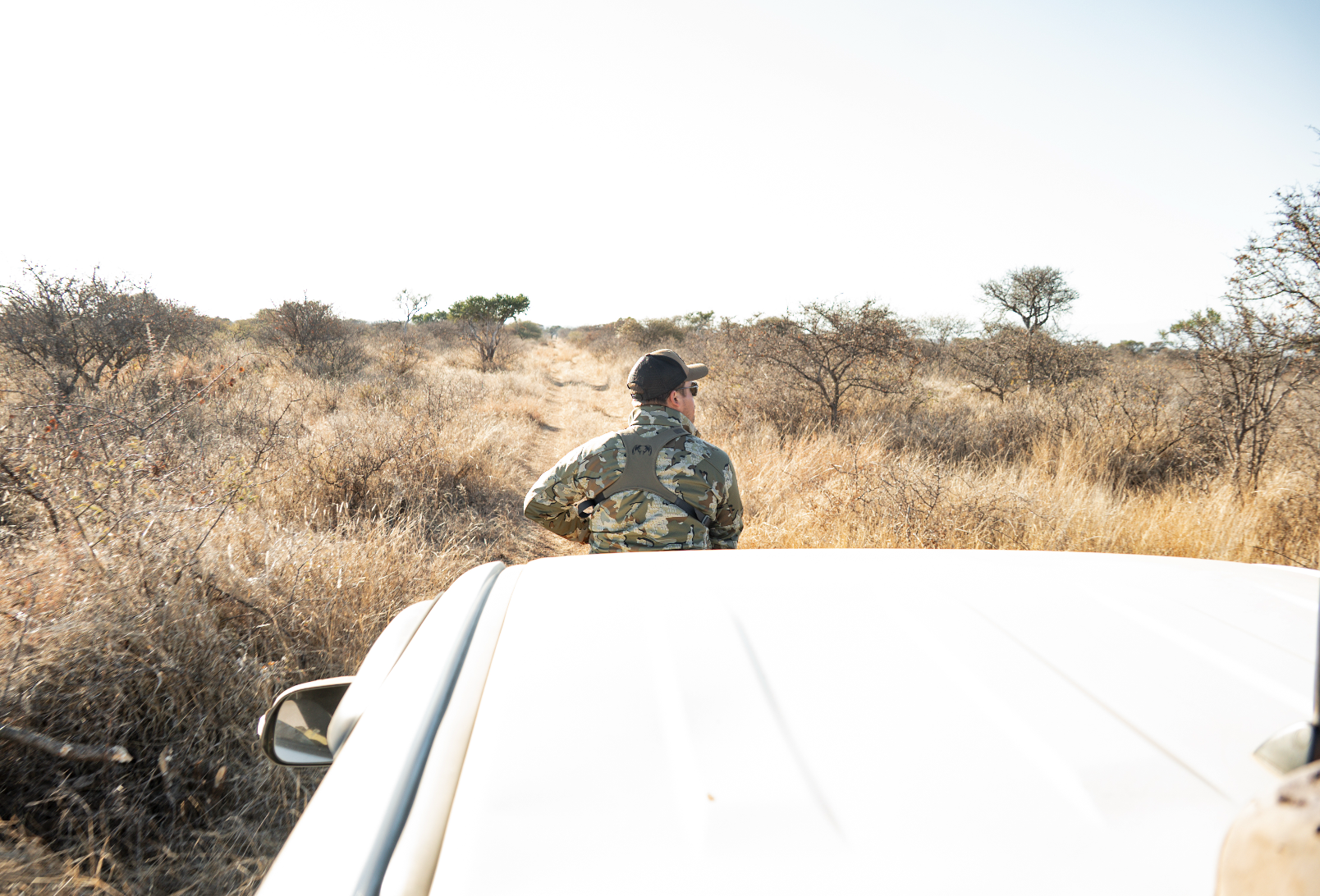 A person in camouflage jacket and cap standing next to a white vehicle in dry, grassy savanna landscape with sparse trees.