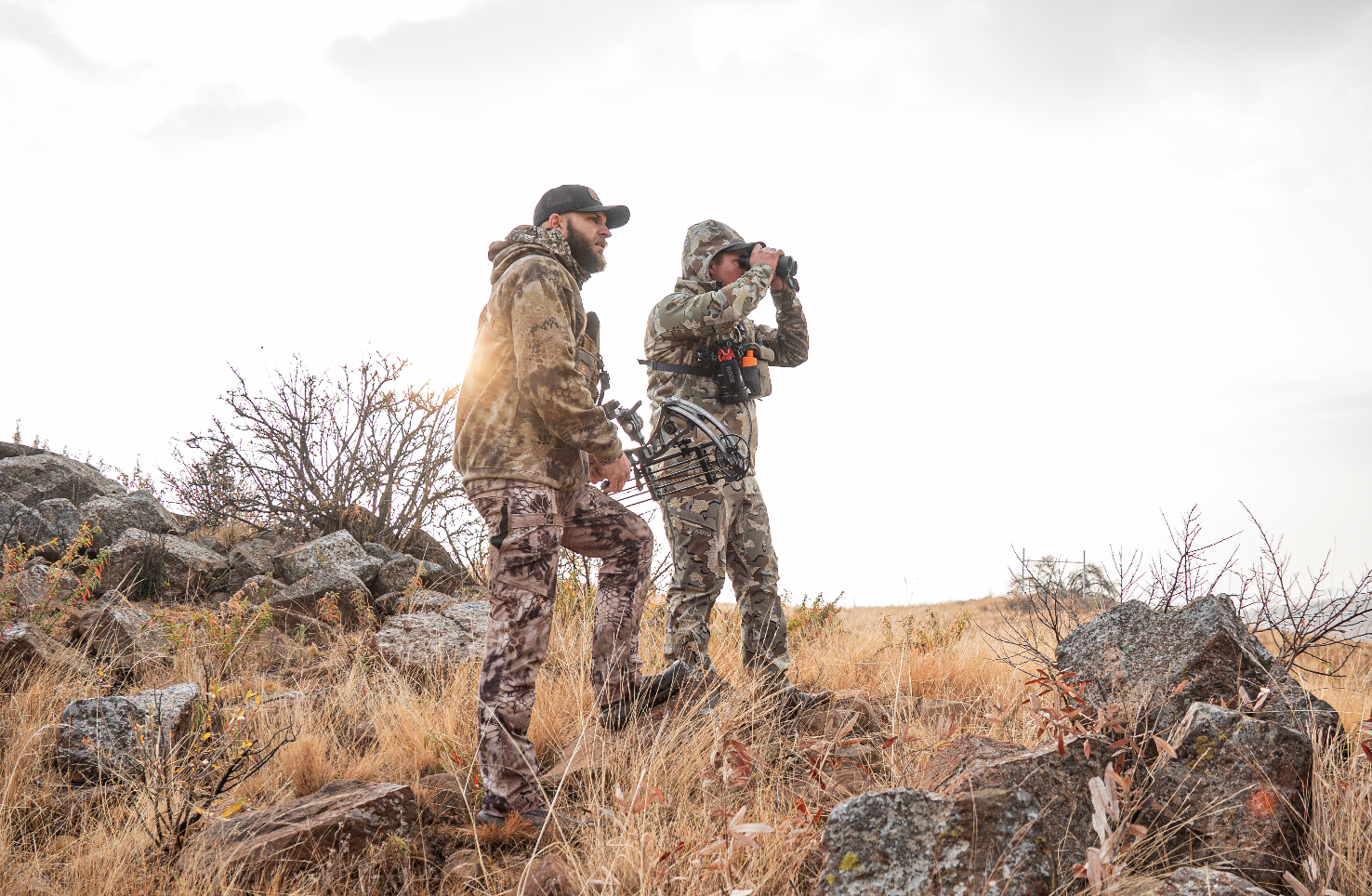 Two men in camouflage clothing exploring a rocky, grassy landscape. One is looking through binoculars while the other stands nearby.