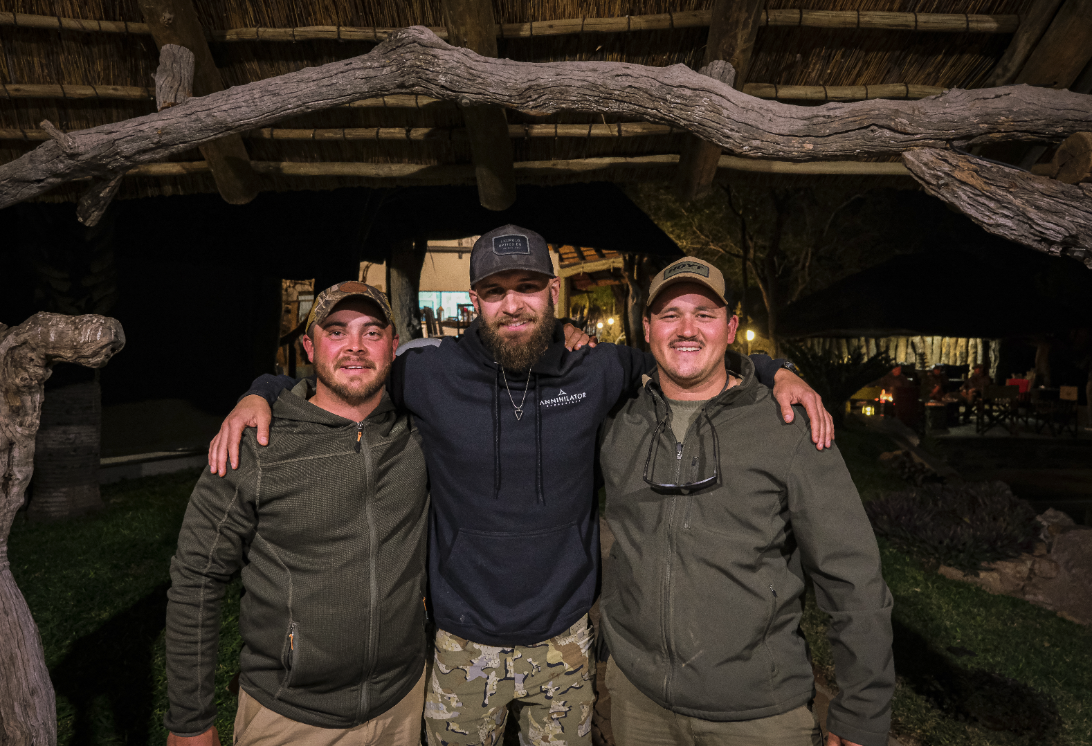 Three men standing outdoors at night, smiling, with their arms around each other, under a rustic wooden archway with a thatched roof.