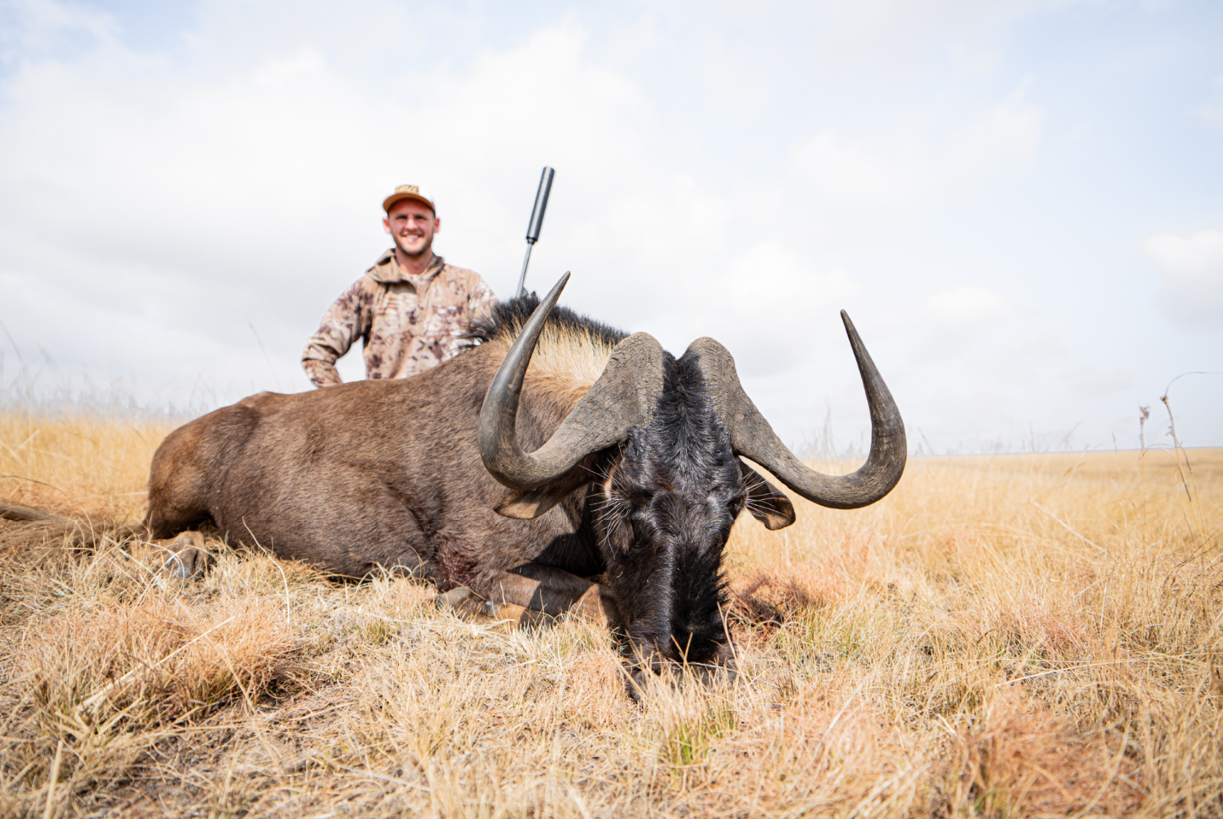 A smiling man in camouflage clothing and a hat standing behind a dead large buffalo with prominent horns, lying in the grassland under a partly cloudy sky.