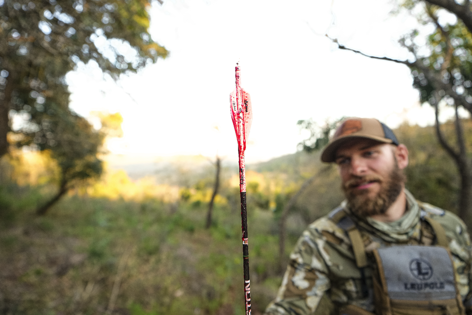 A man with a beard wearing camouflage clothing and a tan cap, sitting outdoors in a wooded area during daylight, with a fishing lure in the foreground.