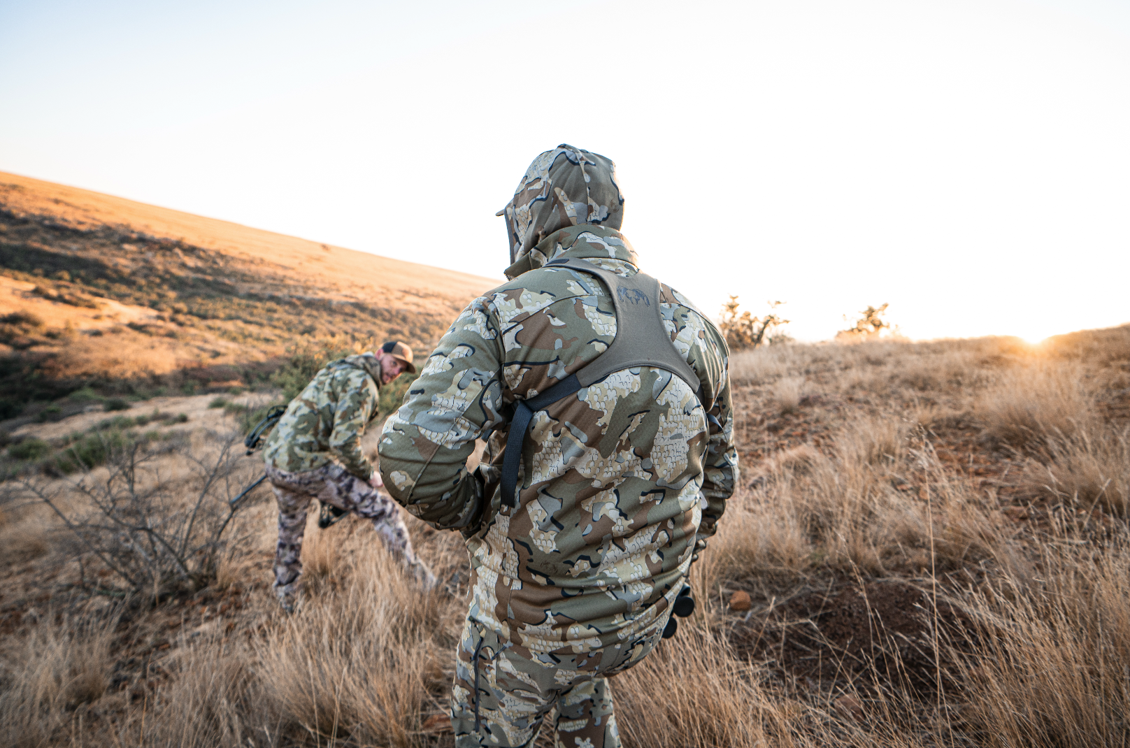 Two people dressed in camouflage hiking gear in a dry, grassy outdoor area at sunset.