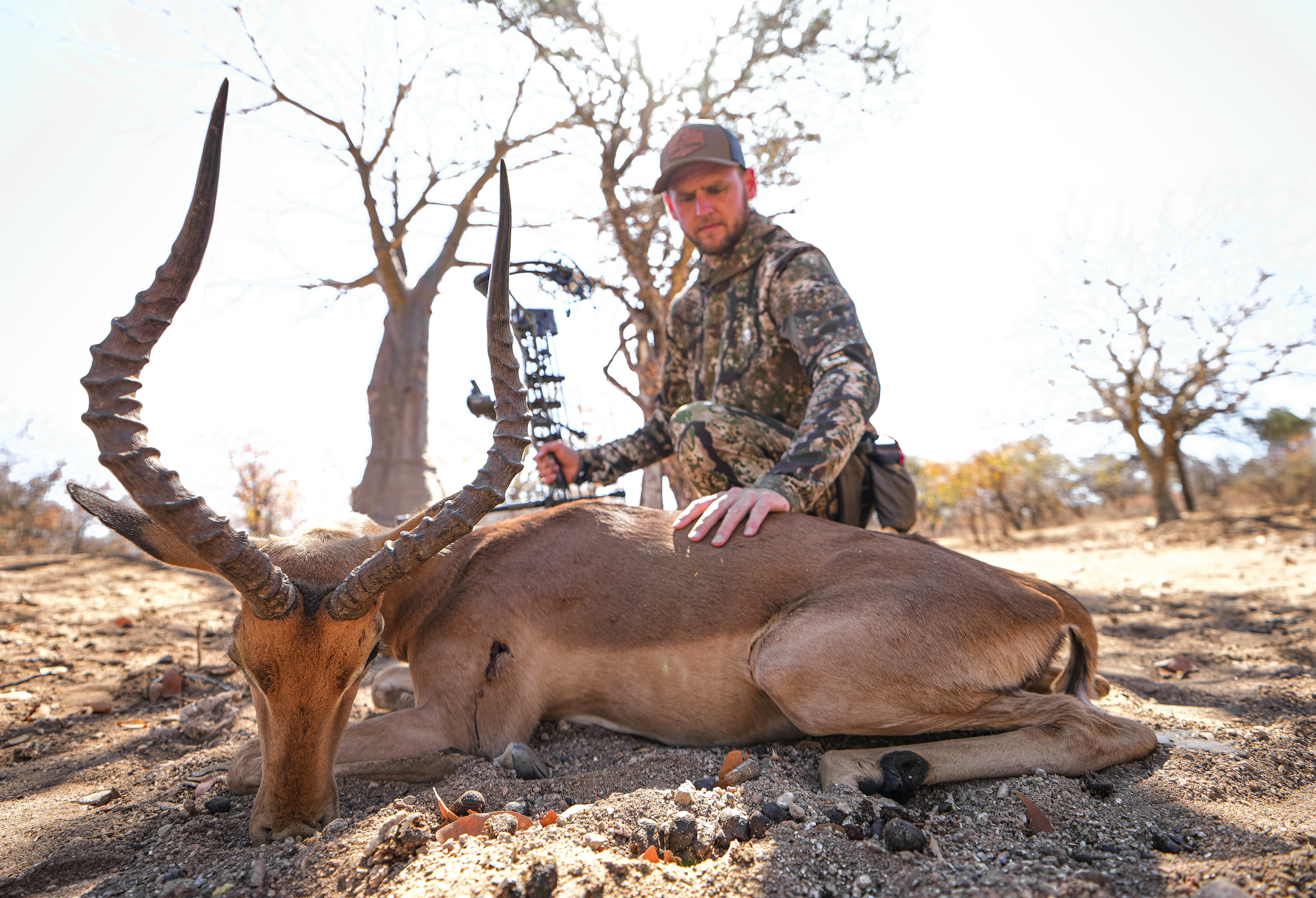 A man in camouflage clothing kneels next to a dead antelope with large, twisted horns, resting on the ground in a dry, open landscape with scattered trees and a bright sky in the background.