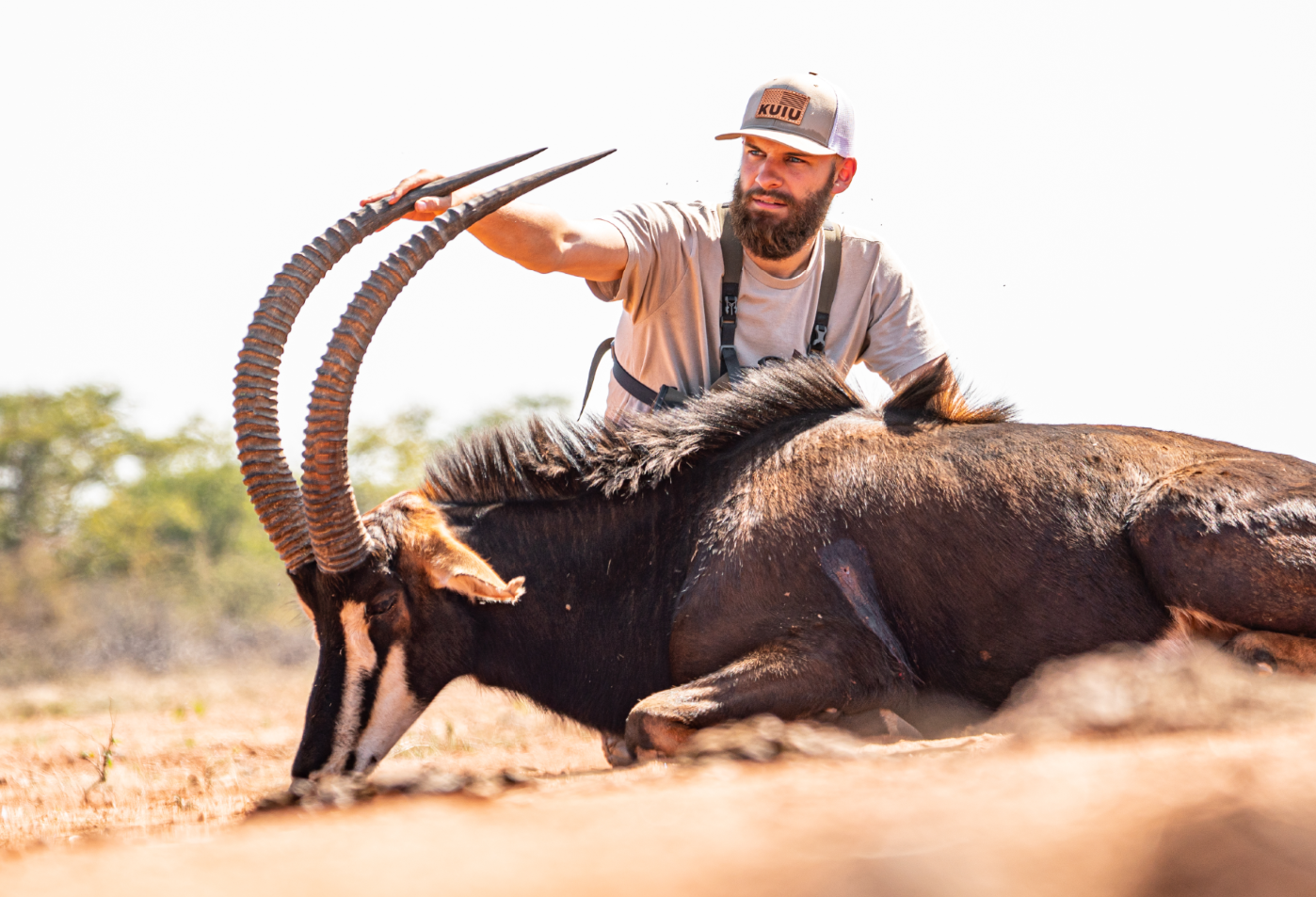 A man with a beard and cap lying on the ground, holding the horns of an antelope with long, curved horns, in a dry, open landscape.