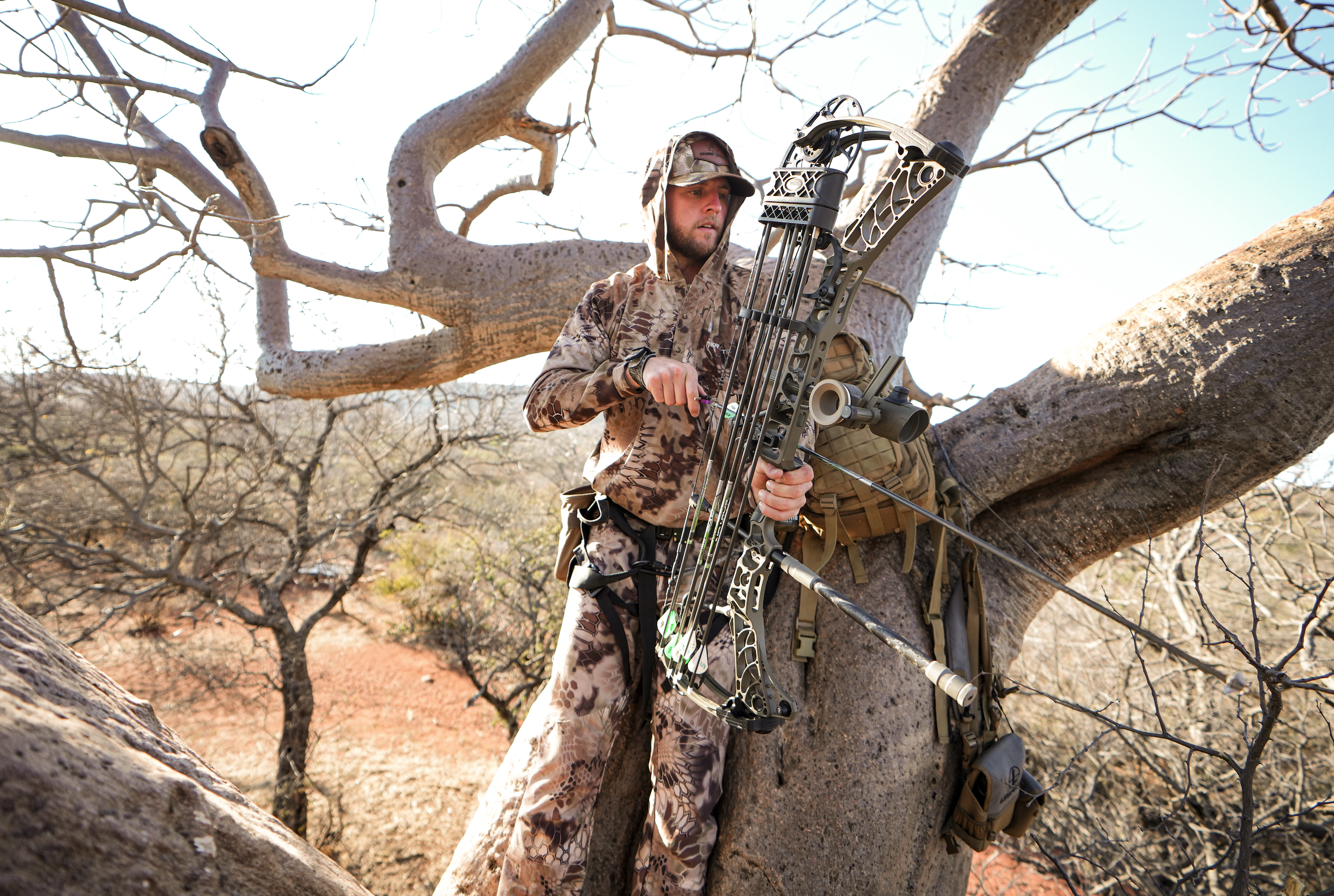 A man in camouflage clothing stands next to a large tree, aiming a compound bow with arrows, in a desert landscape with bare trees.