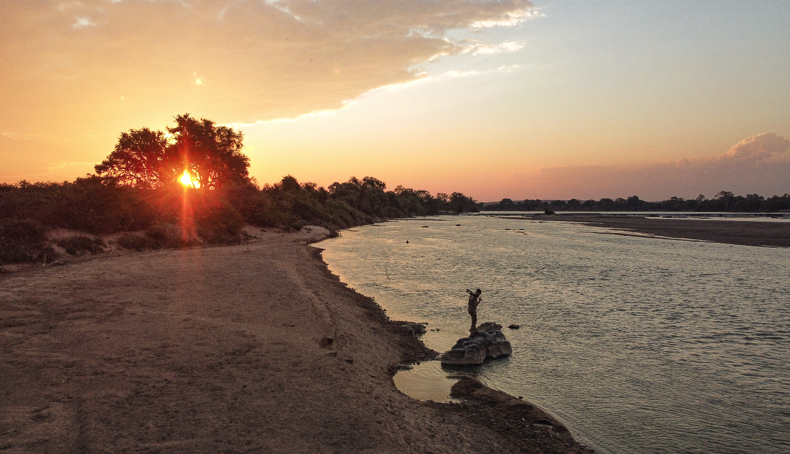 A person standing on a rock at the edge of a river during sunset, with the sun partially hidden behind trees on the left side of the image. The sky is partly cloudy with warm hues, and the river extends into the distance with trees lining the far ban