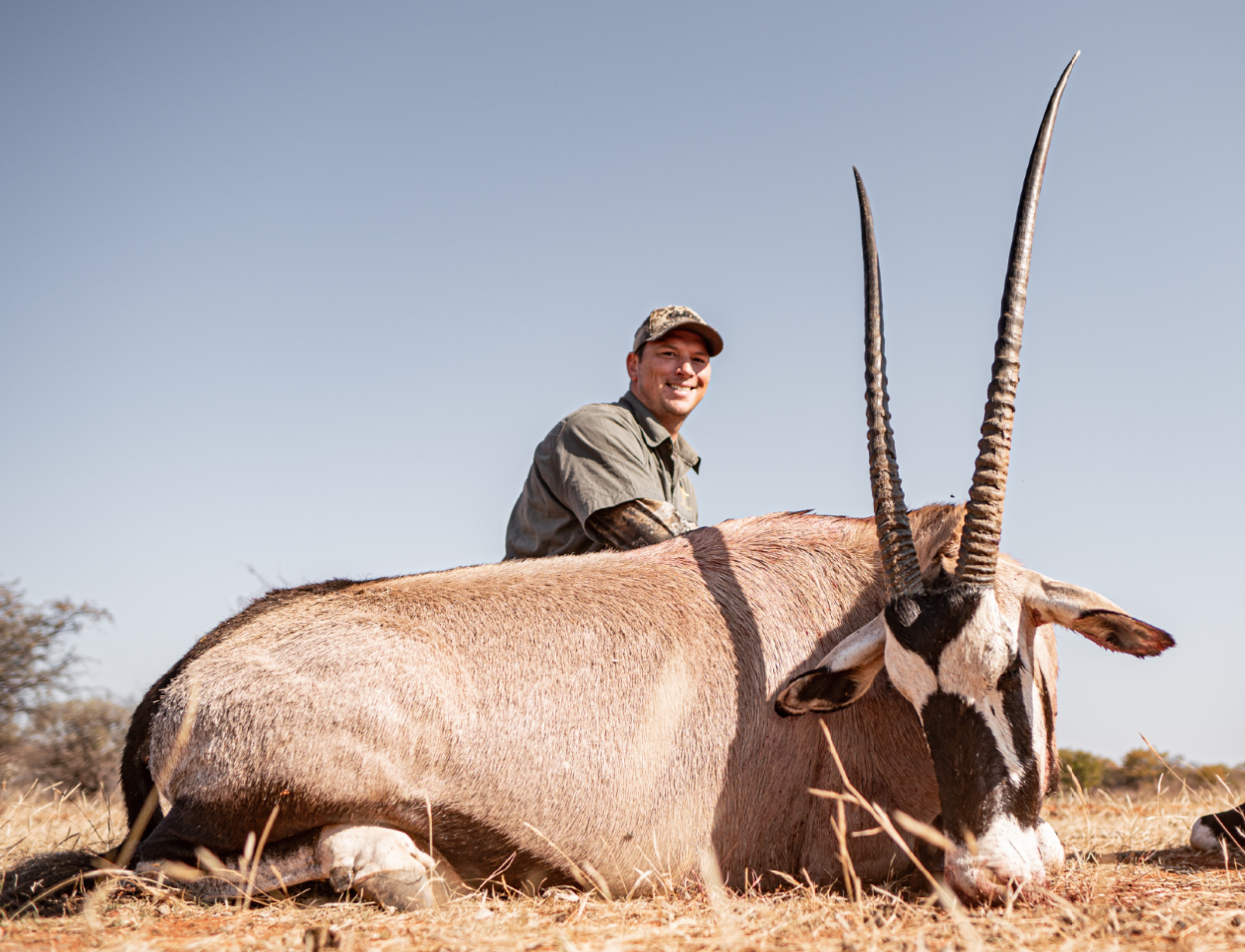 A man smiling and sitting next to a large dead oryx with long, straight horns lying on dry grass in an open landscape.