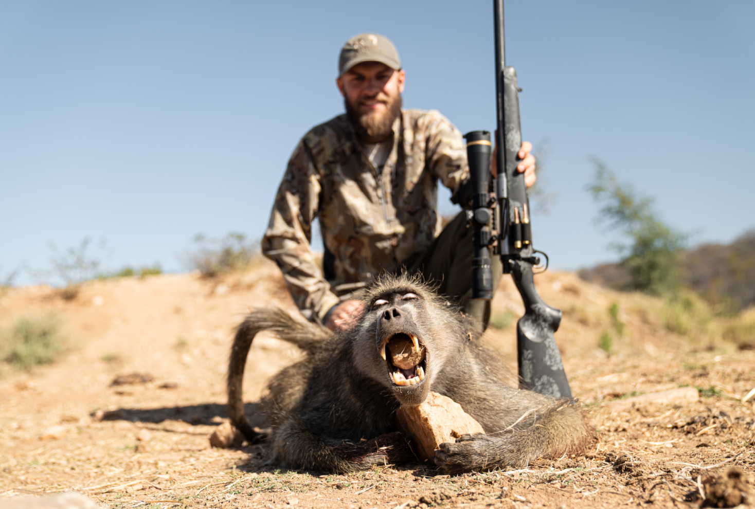 A man in camouflage clothing with a hunting rifle kneels next to a dead baboon on the ground, with its mouth open in a roar. The background is a dry, open landscape with bushes and a clear blue sky.