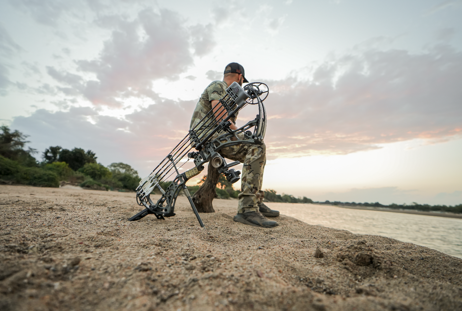 A person in camouflage clothing sitting on a sandy riverbank with a compound bow leaning against the ground near them, under a sky with scattered clouds during sunset.