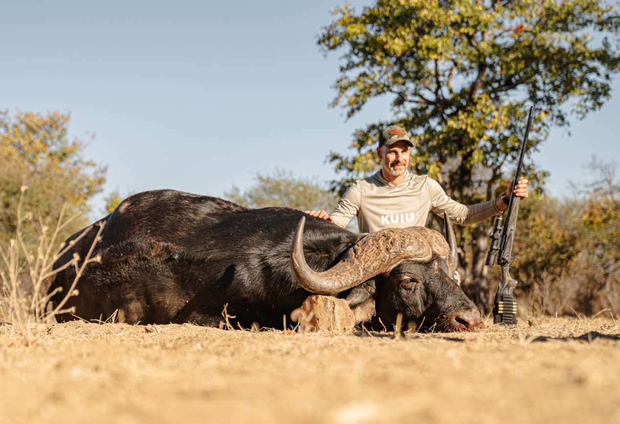 A man kneeling next to a large, dead buffalo on the ground in a dry, grassy area with trees in the background. The man is holding a rifle and smiling, wearing a beige long-sleeve shirt and a camouflage cap.