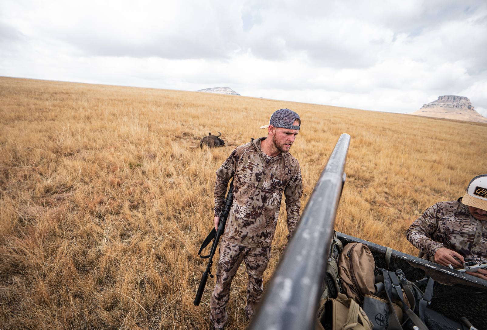 Man in camouflage clothing and a baseball cap holding a rifle, standing in a grassy field, with another person on the right looking at a phone, and a buffalo lying in the background.