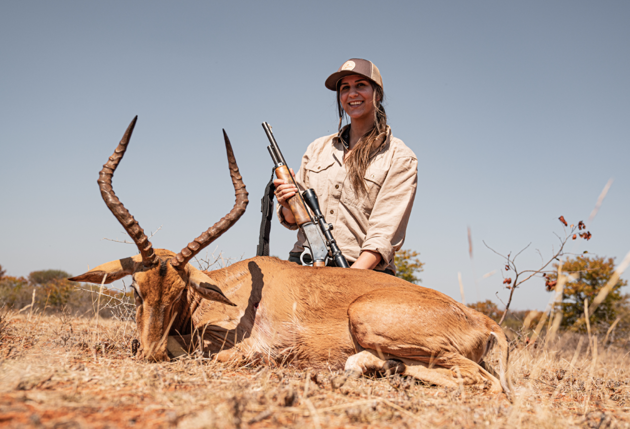 A woman with long hair, wearing a beige shirt and baseball cap, smiling and holding a rifle, kneeling next to a large antelope with spiraled horns lying on the ground in a dry open field under clear skies.
