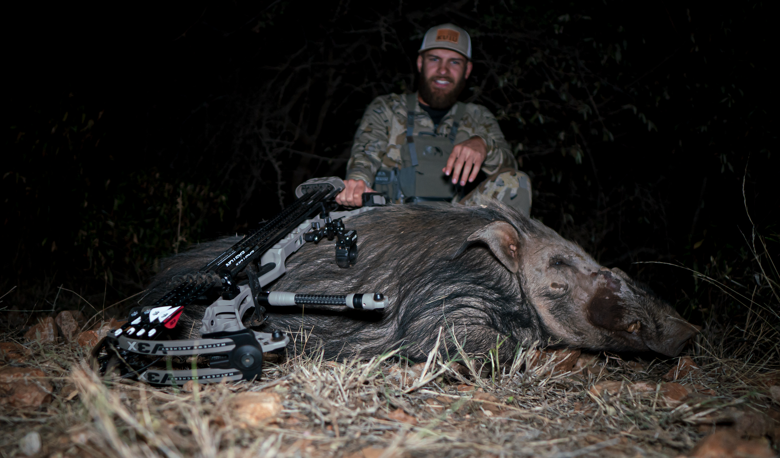 A hunter crouching behind a large dead pig with a bow and arrow lying on the ground in front of the pig, at night in a wooded area.