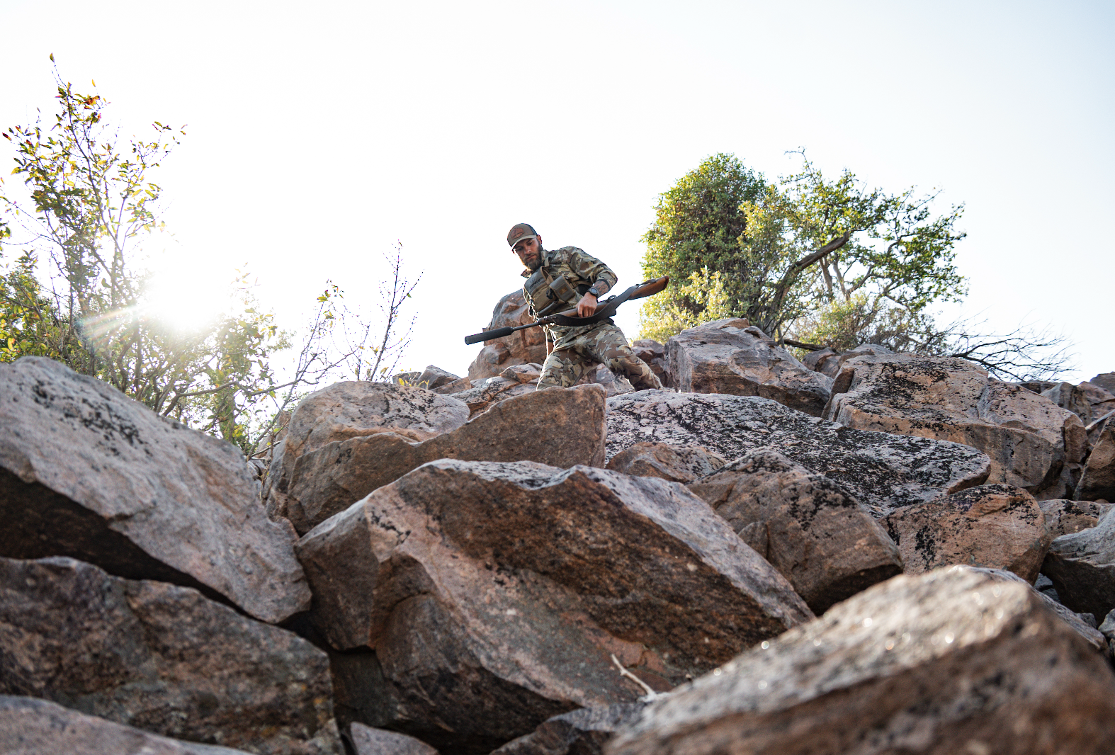 A soldier in camouflage uniform carrying a shovel climbs over large rocks outdoors with trees and clear sky in the background.