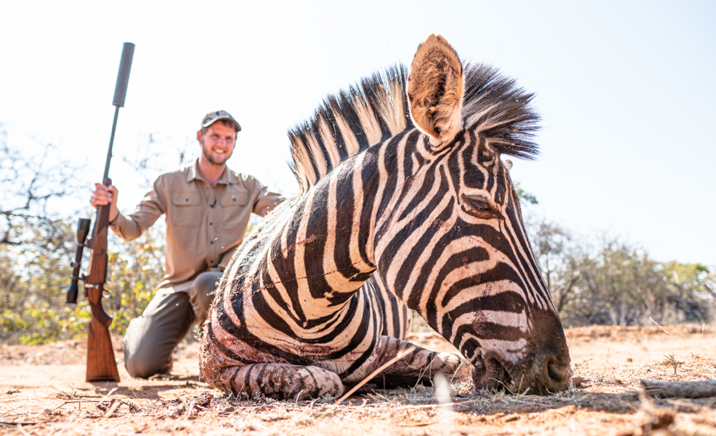 A smiling man kneeling behind a lying zebra in a dry, open landscape.