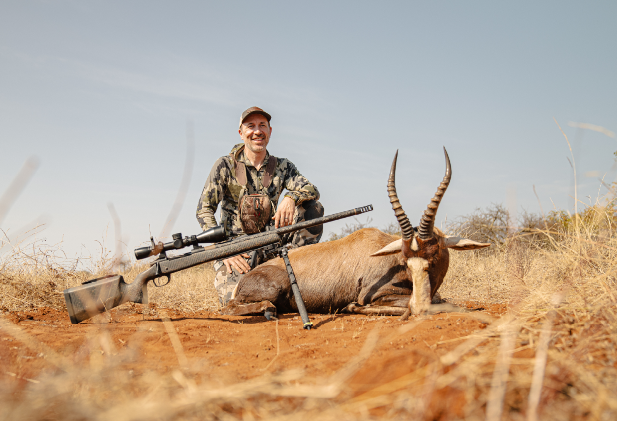 Man in camouflage hunting gear kneeling next to a lying kudu with twisted horns in an open dry landscape, holding a rifle.