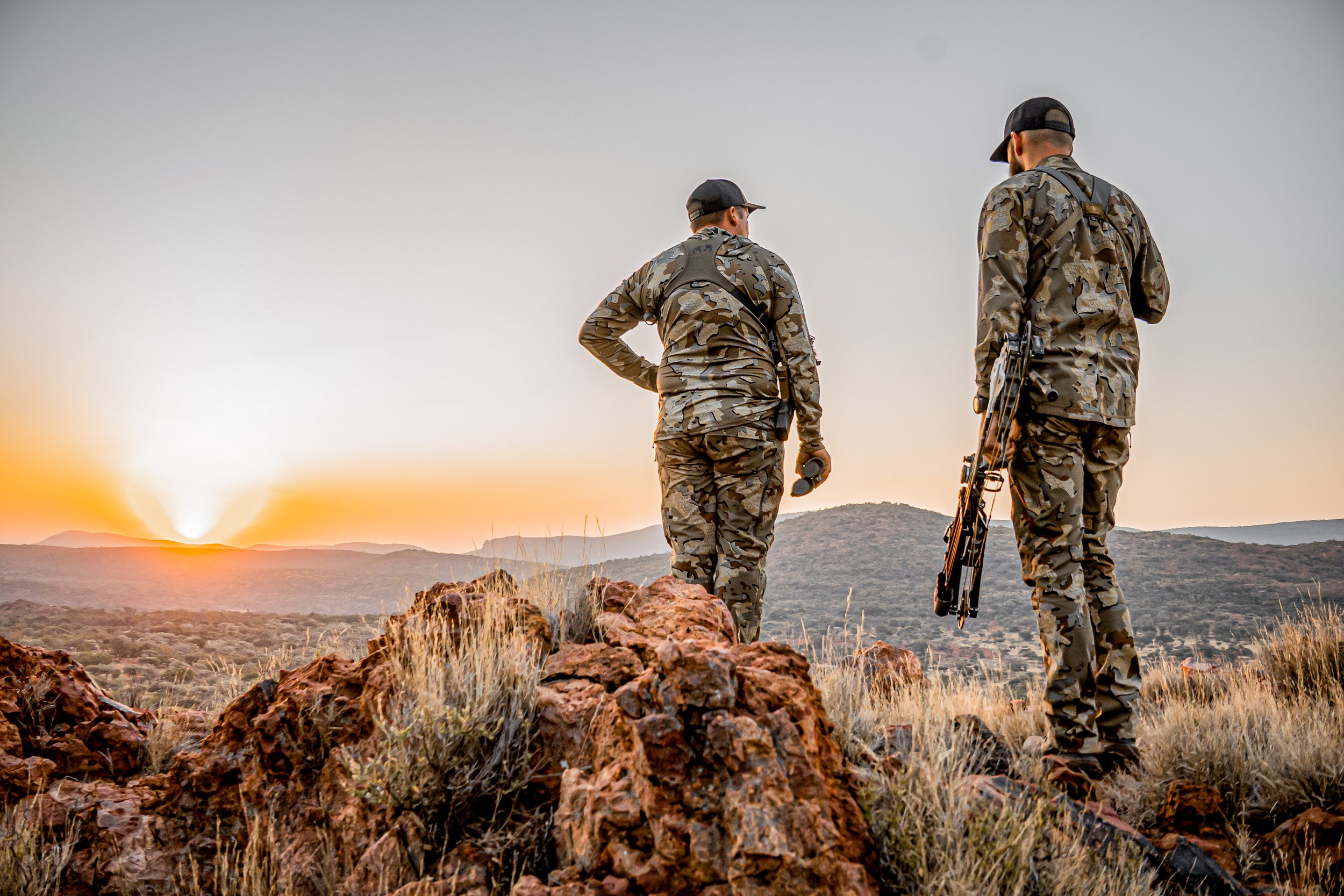 Two soldiers in camouflage uniform standing on rocky terrain during sunset, overlooking a mountainous landscape.