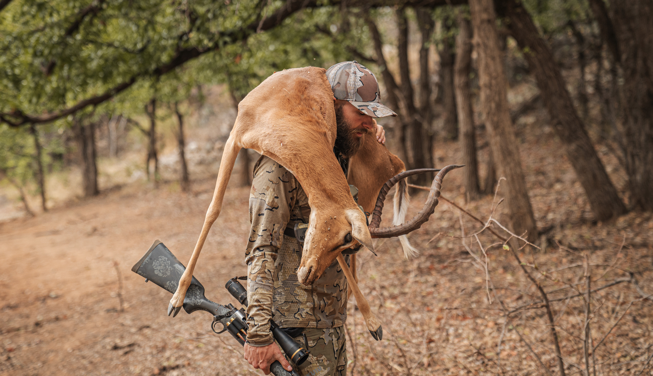 A hunter in camouflage clothing carrying a rifle with a mounted scope, holding a large antelope with long curved horns over his shoulder in a wooded area.