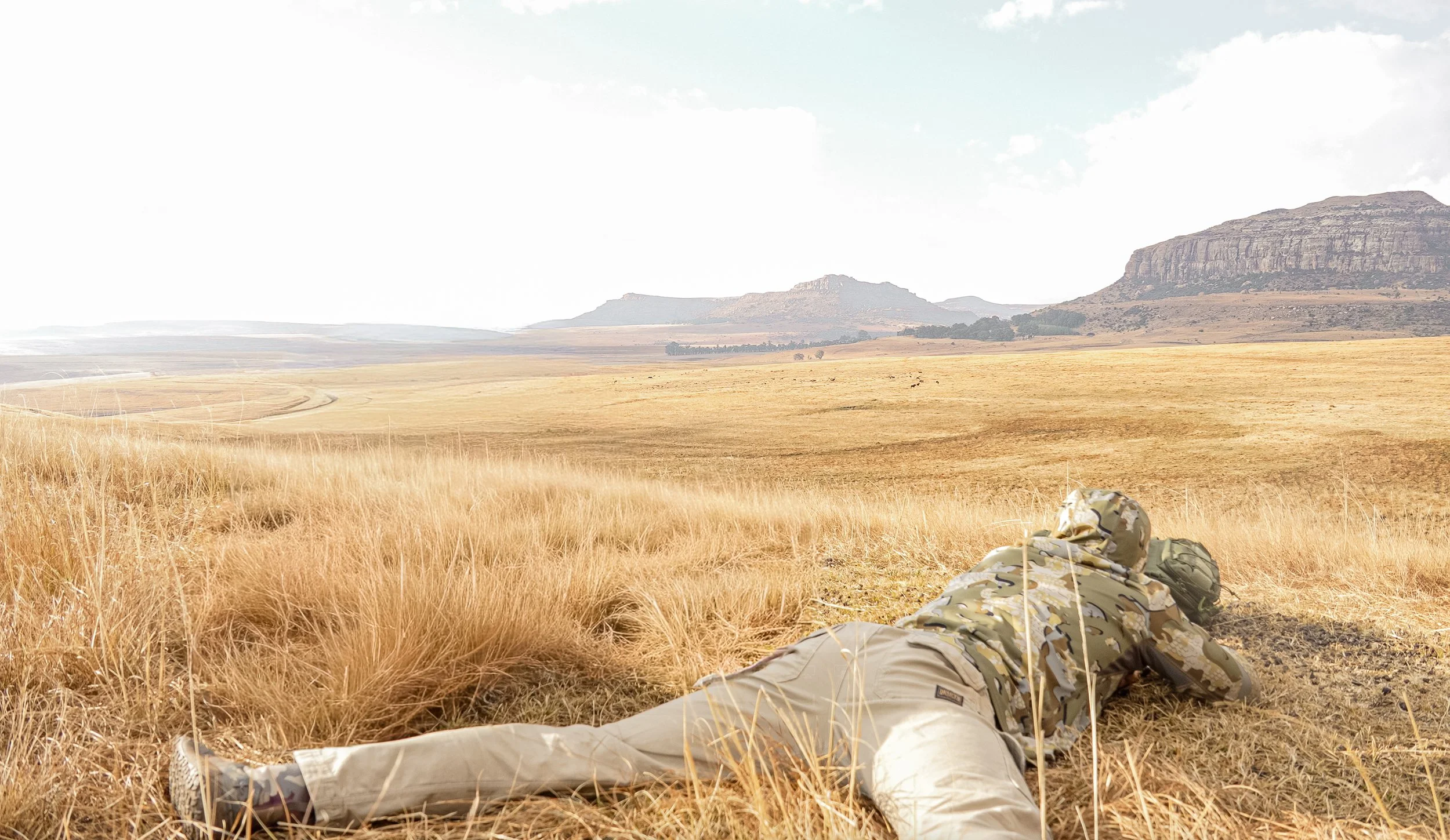 A soldier lying prone in a dry, grassy plain with mountains and a blue sky in the background.