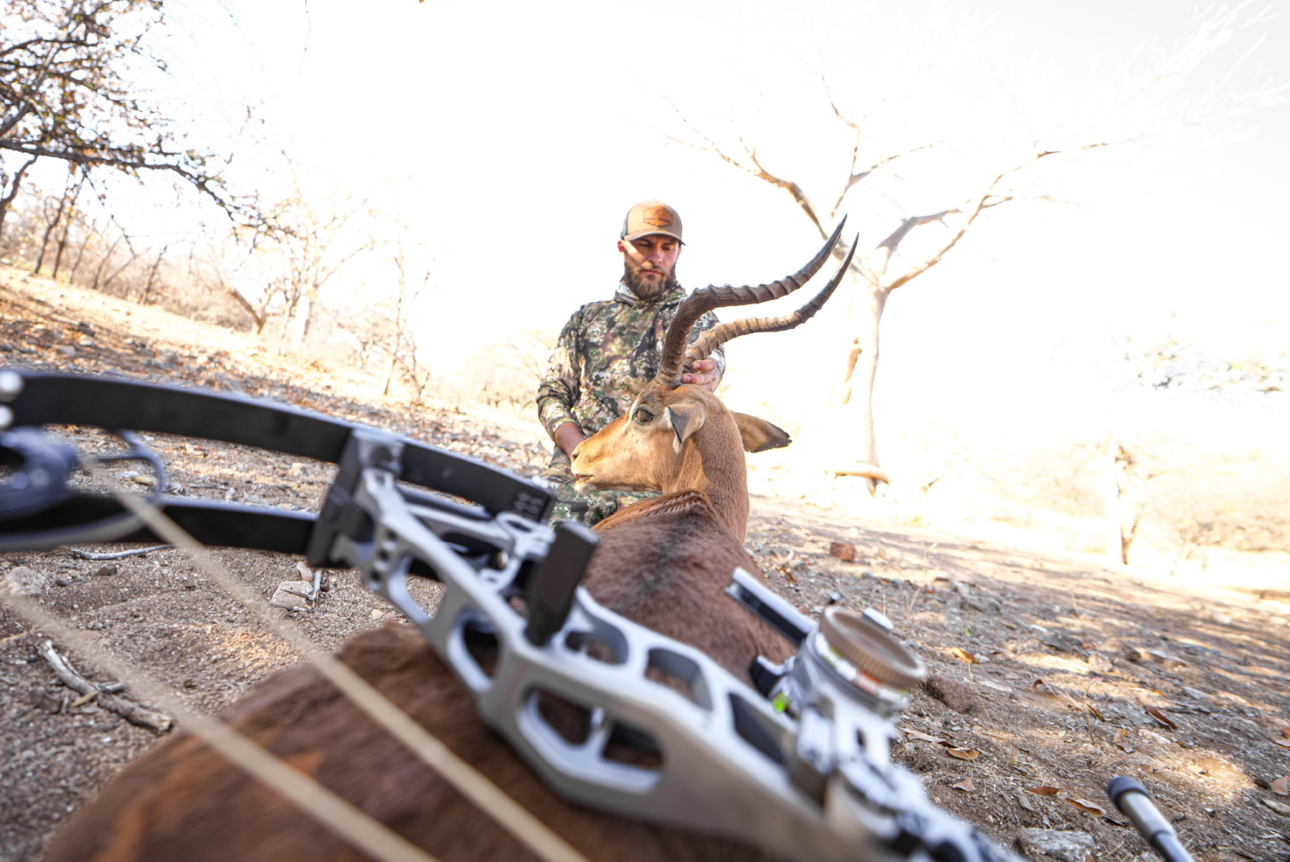 A man in camouflage clothing and a cap stands outdoors holding a large antelope with long, curved horns. A modern compound bow is lying on the ground in the foreground, indicating a hunting scene. The background features leafless trees and a dry, ope