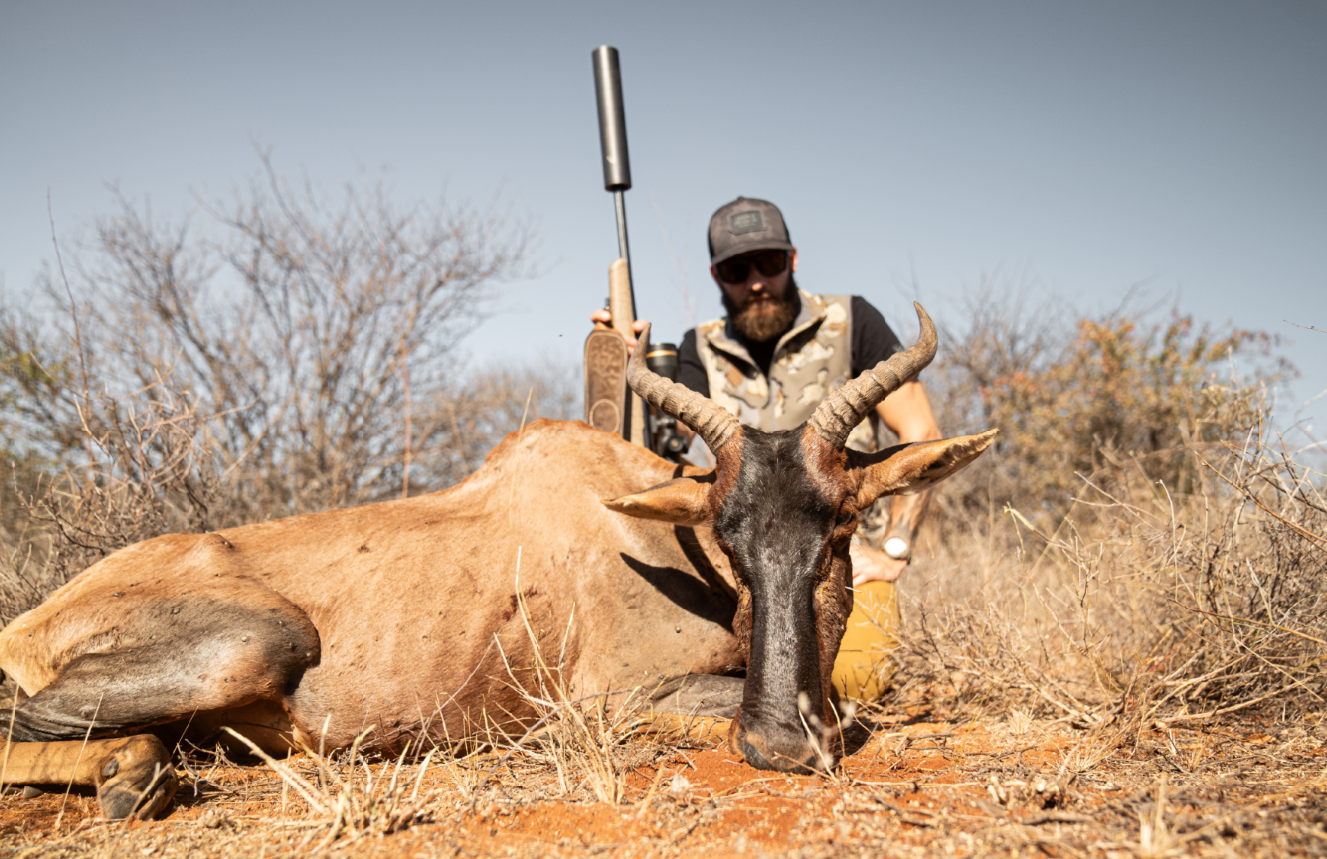 A man with a beard, wearing sunglasses, a cap, and a camouflage vest, kneels behind a dead antelope with large curved horns in a dry, bushy landscape. The man holds a rifle upright behind his head.