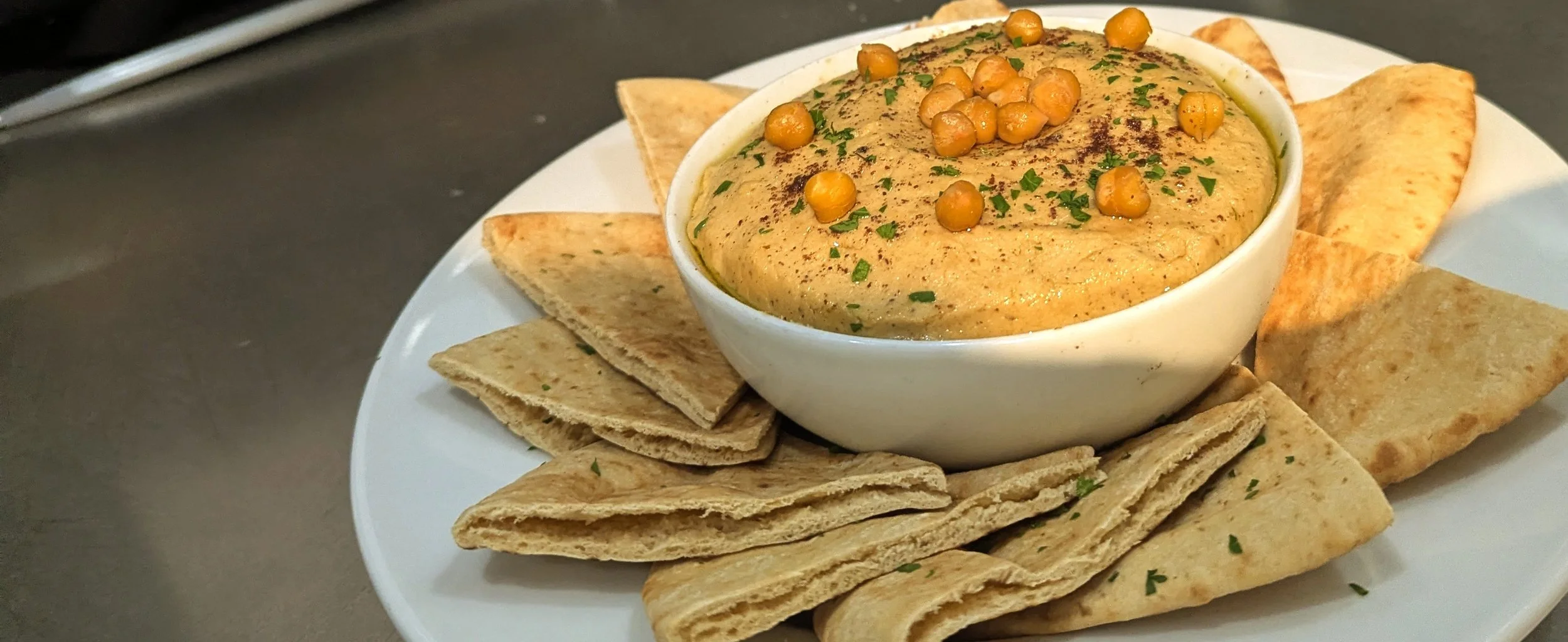 A white bowl filled with creamy hummus topped with whole chickpeas and chopped parsley, surrounded by pita chips on a white plate.