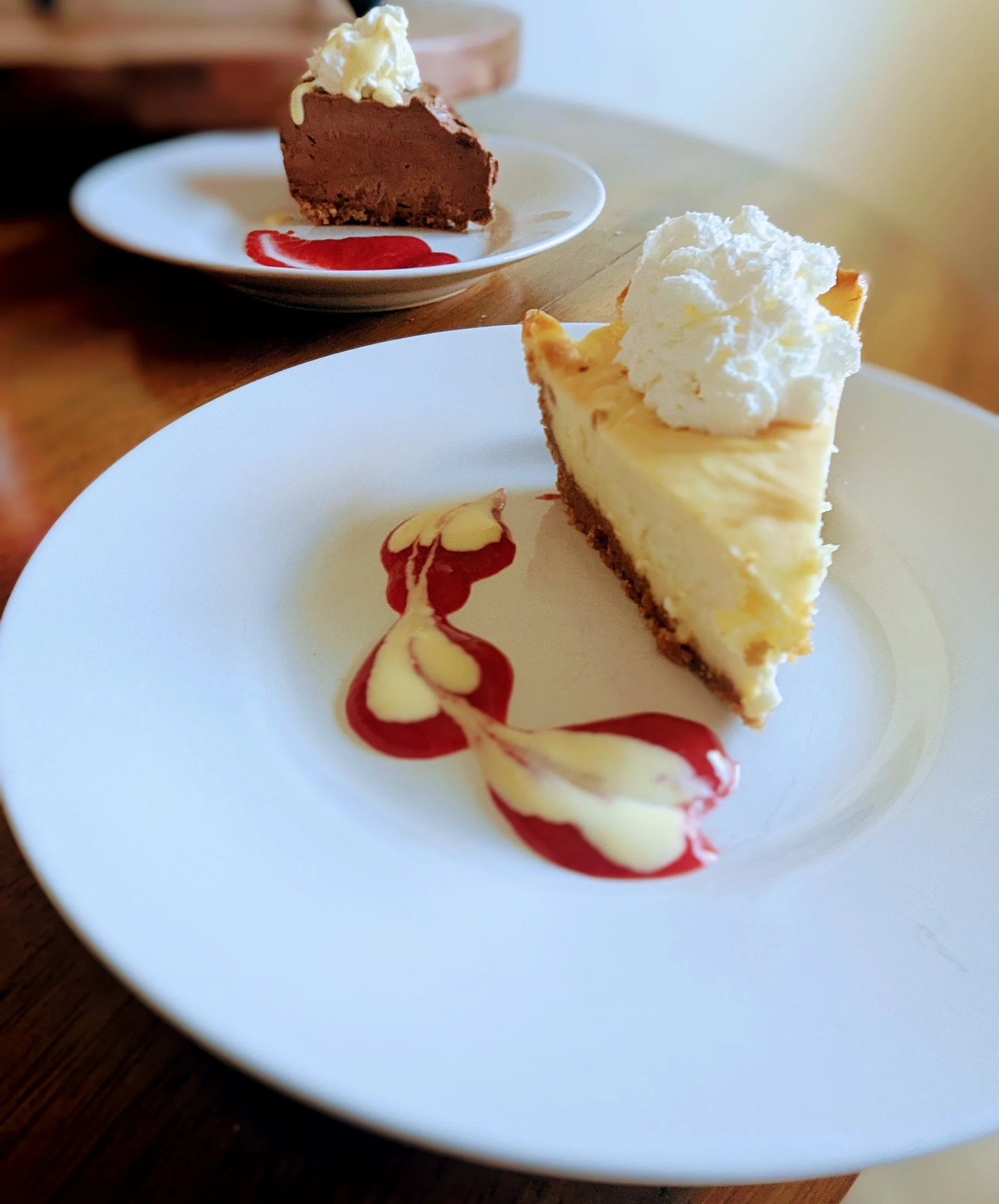Two slices of cheesecake with whipped cream and raspberry sauce on white plates, one in the foreground with a decorative raspberry sauce design, and the other in the background with a chocolate mousse cake topped with whipped cream.